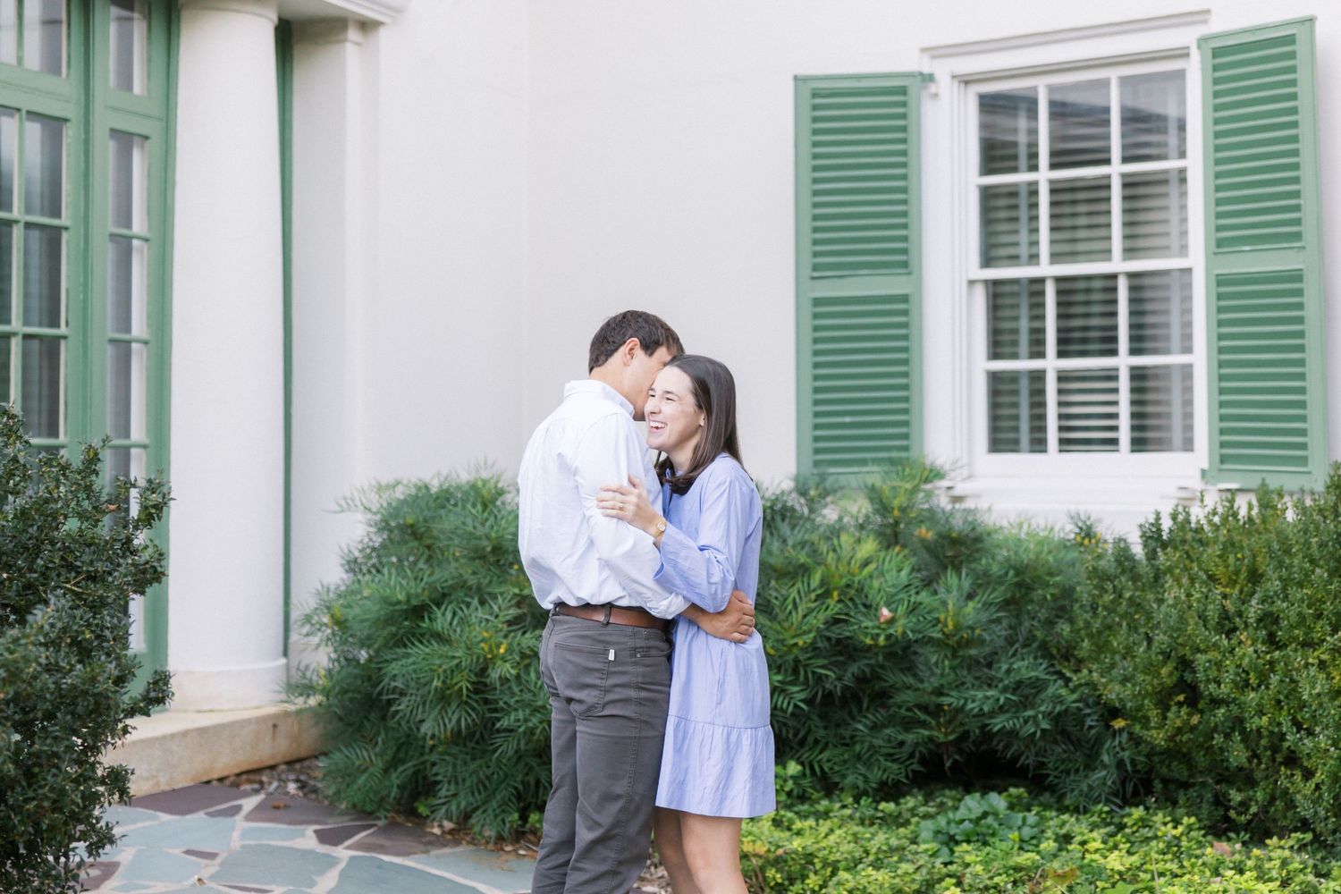 A couple shares a tender embrace outside a white building with green shutters and lush garden landscaping.