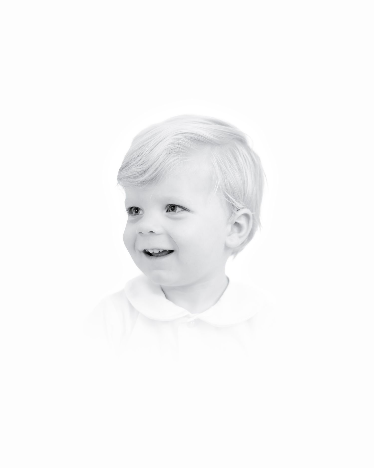 Black and white portrait of a young child with light hair smiling warmly against a white background.