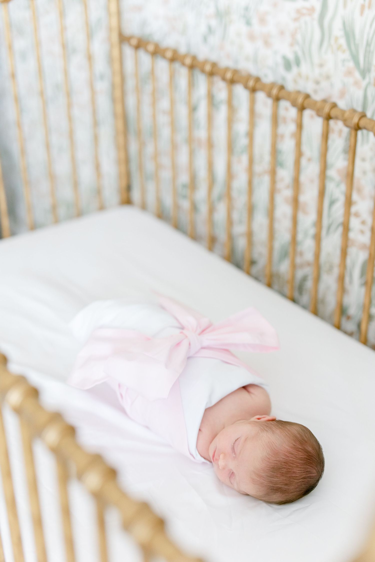 Newborn baby sleeping peacefully in gold crib with pink bow and white bedding.