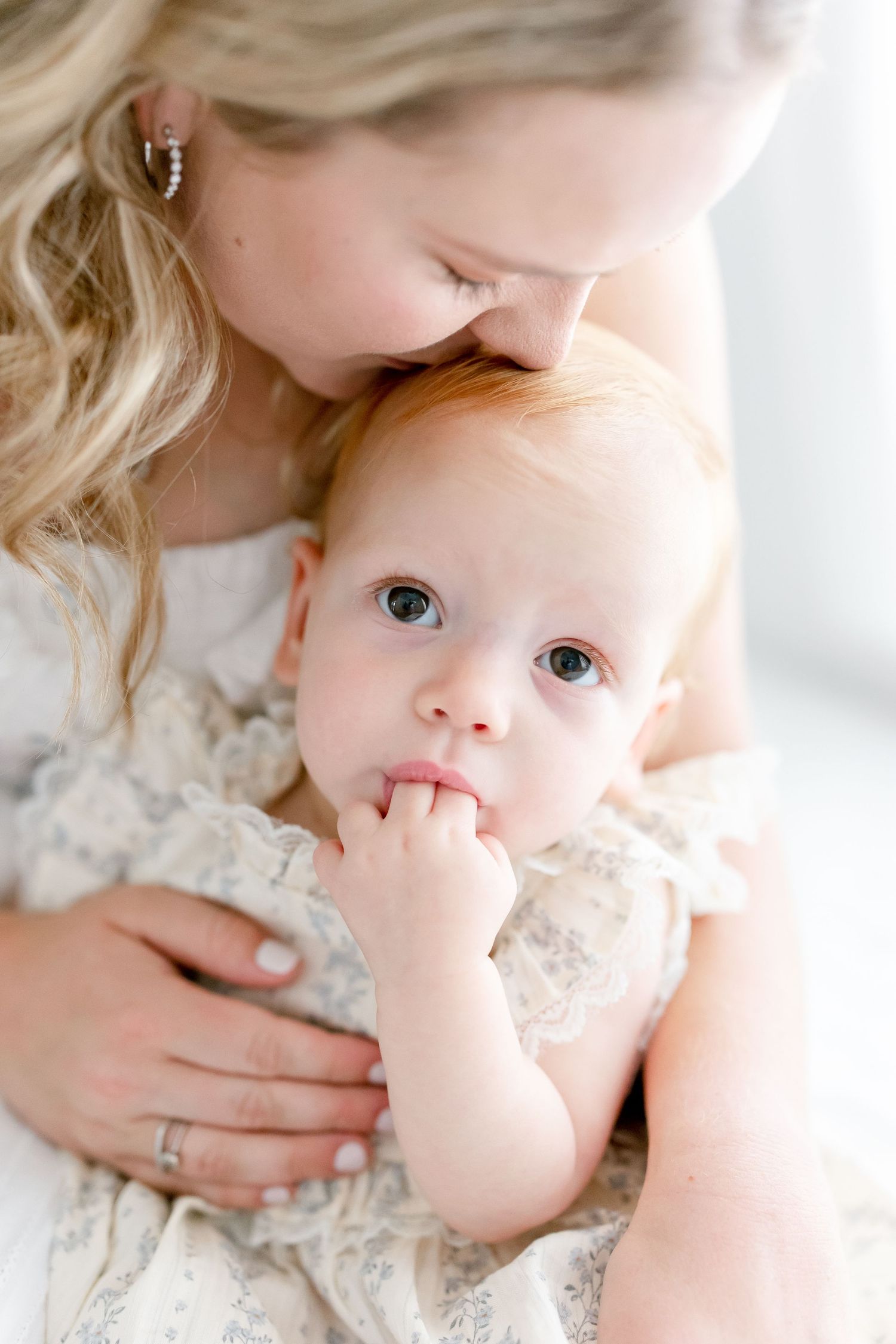 Baby with big bright eyes wearing a lace dress looks up while being held close in a tender maternal embrace.