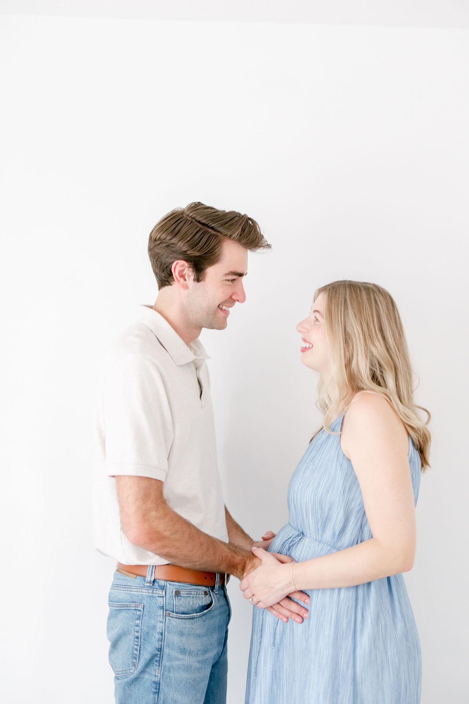 Expecting couple in white and blue outfits share playful moment against white studio background.