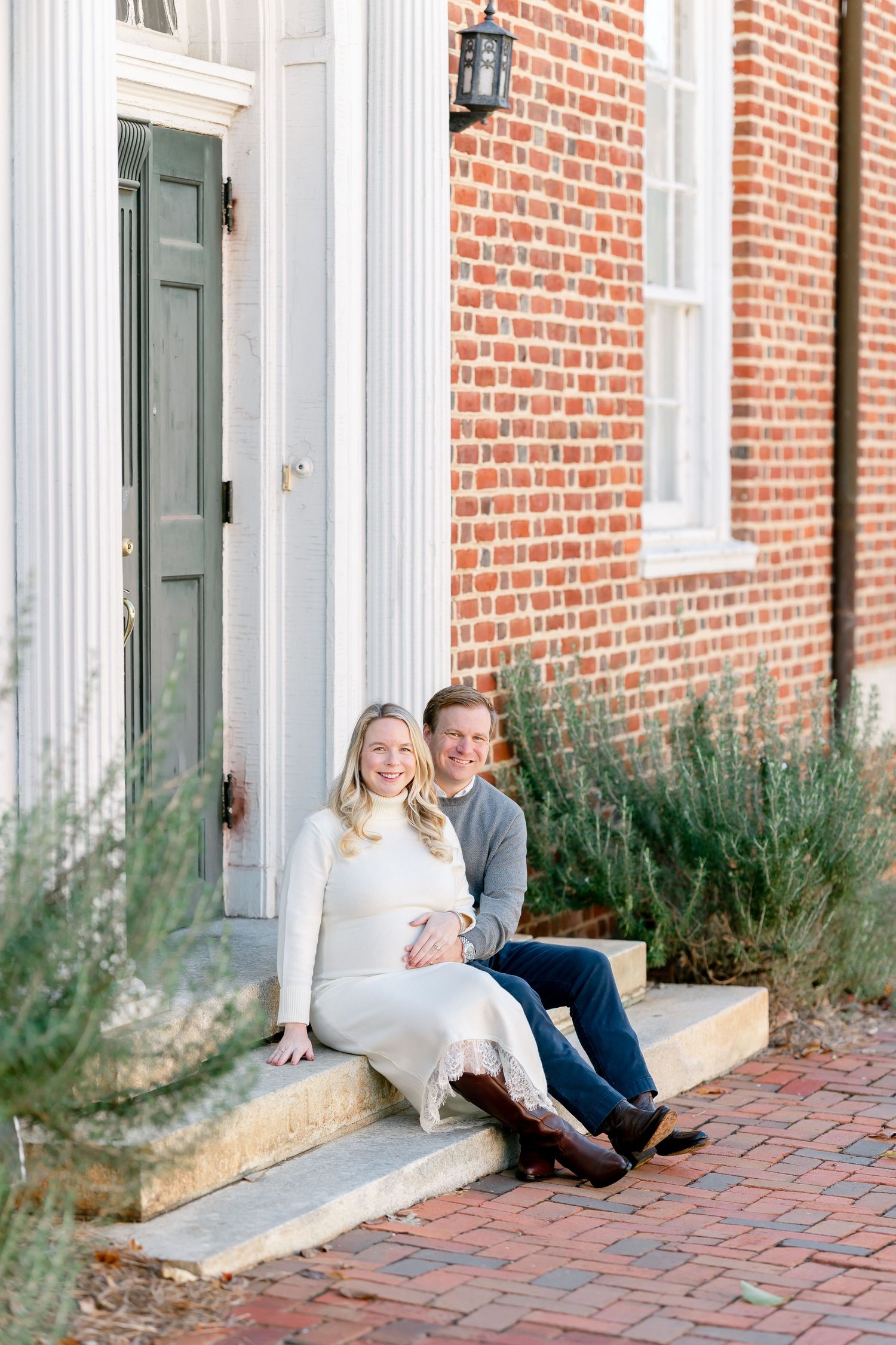 Smiling couple sits together on stone steps outside a historic red brick building with a green door, the woman in a cream dress and boots.