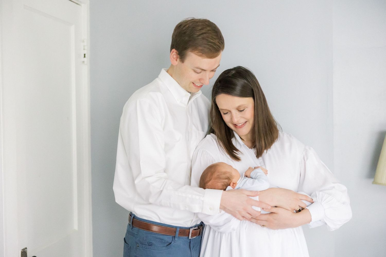 A couple in white clothing tenderly holds and gazes at their newborn baby against a light gray wall.