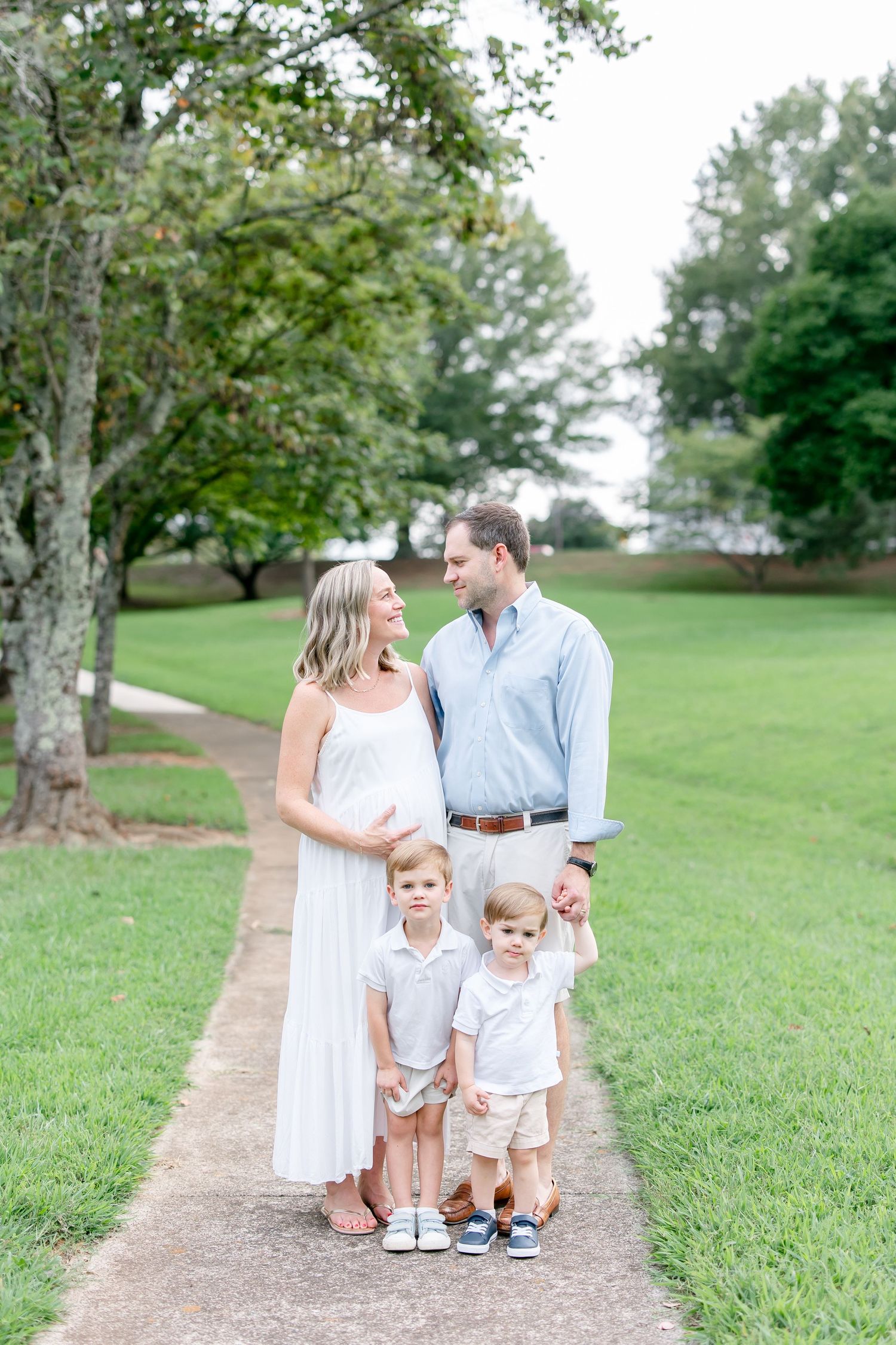 A family of four in white and blue outfits standing on a dirt path surrounded by green grass in a park setting.