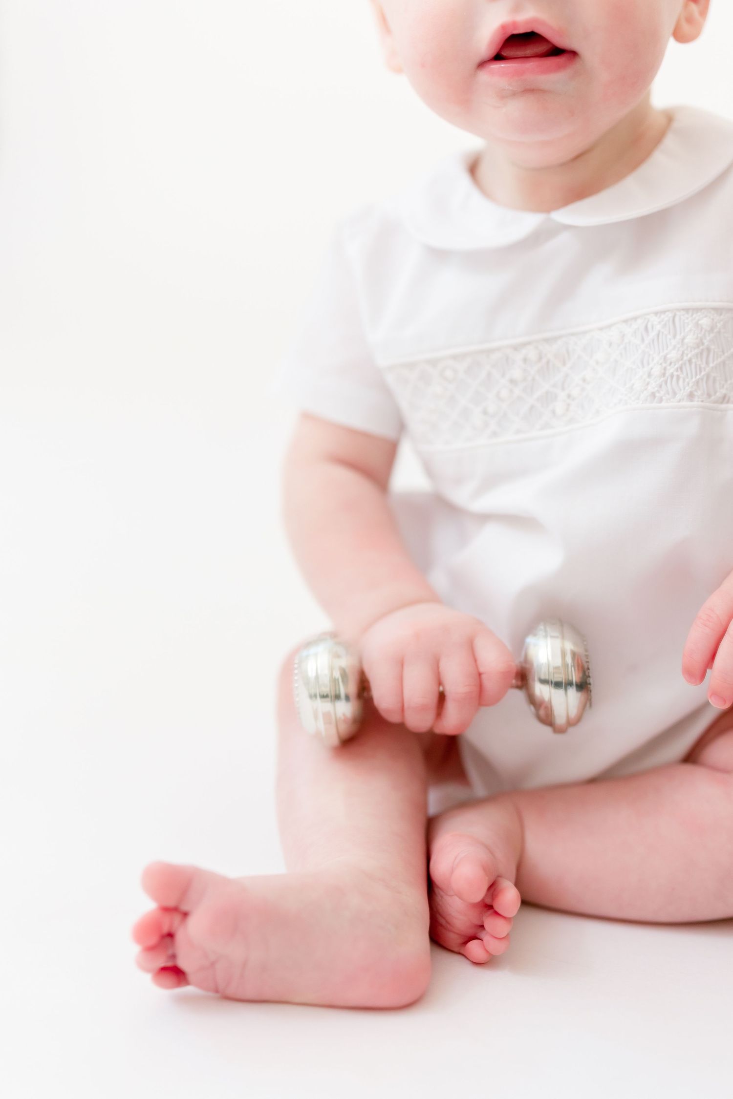 Baby in a white smocked collared romper sits barefoot on a white background, holding a silver dumbbell-shaped rattle.