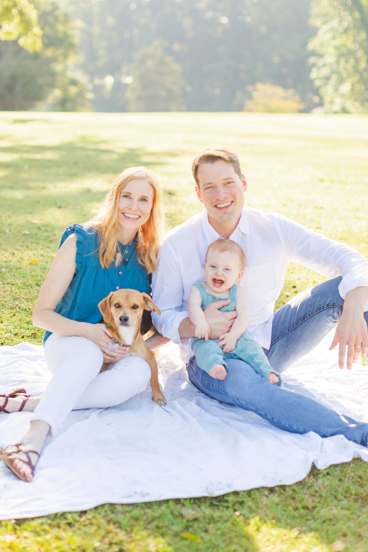 Family enjoying a sunny day outdoors on white blanket with their brown dog in a park setting.