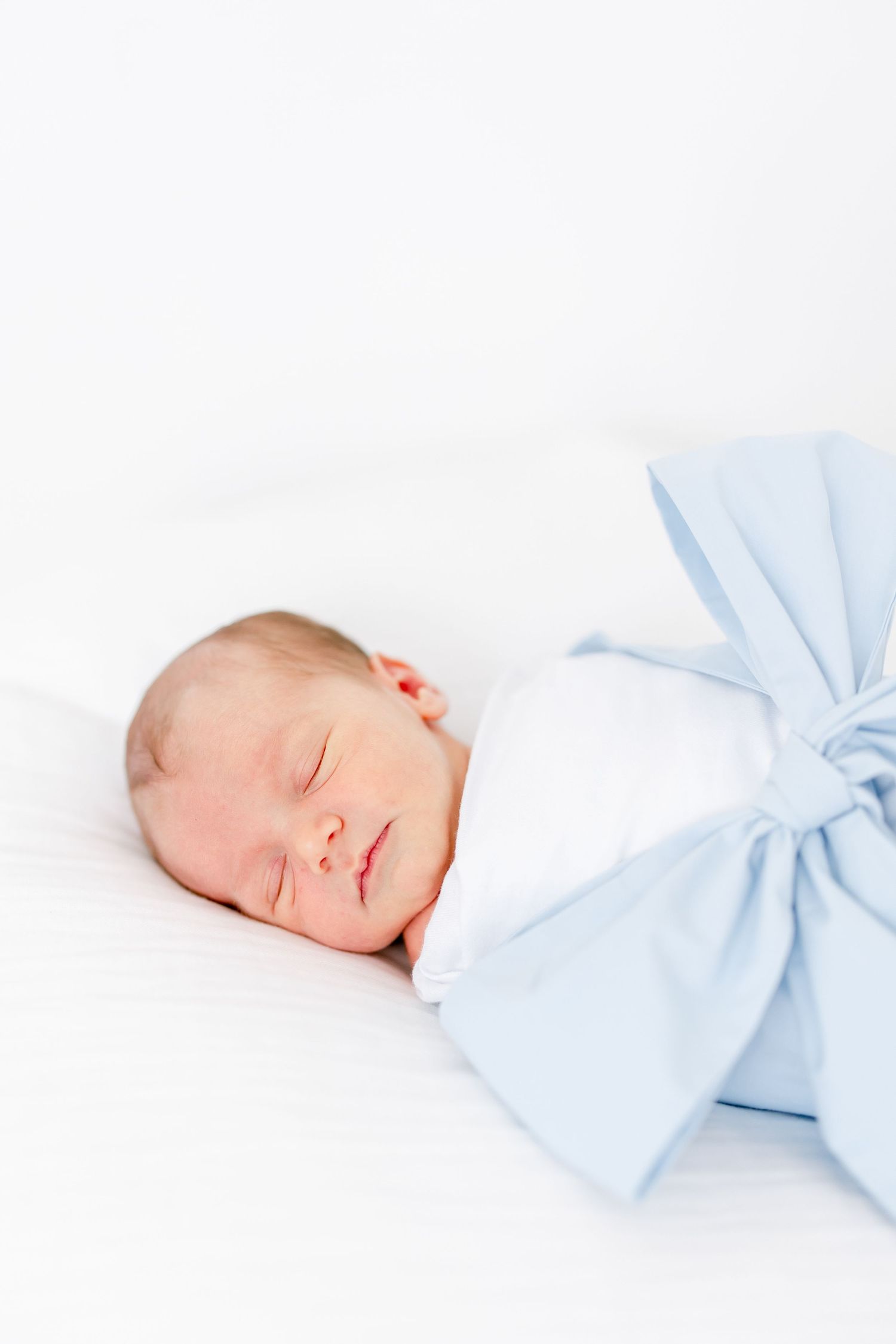 A sleeping newborn baby wrapped in a light blue bow blanket rests peacefully on white bedding.