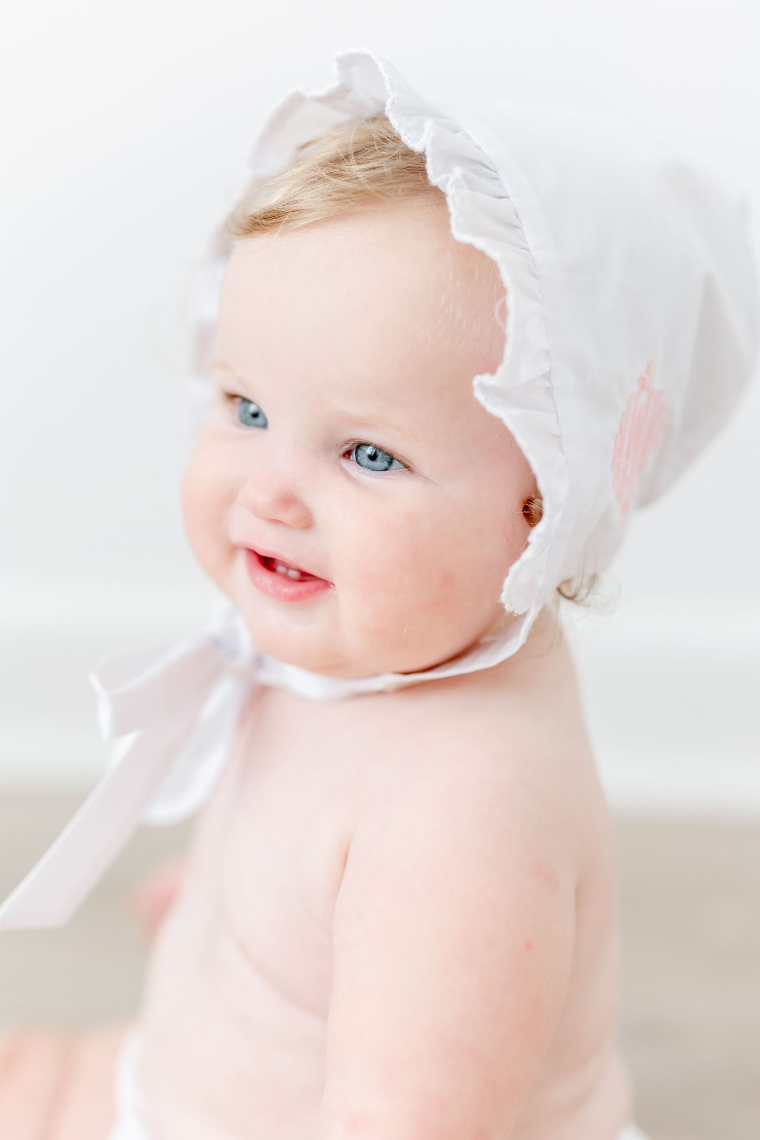 A baby wearing a delicate white bonnet smiles softly in a bright, high-key portrait series.