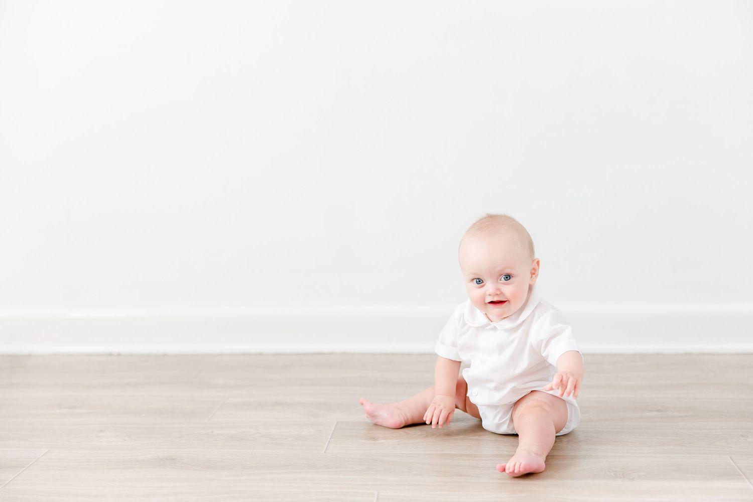 Baby in white onesie sits on light wooden floor against neutral wall background.