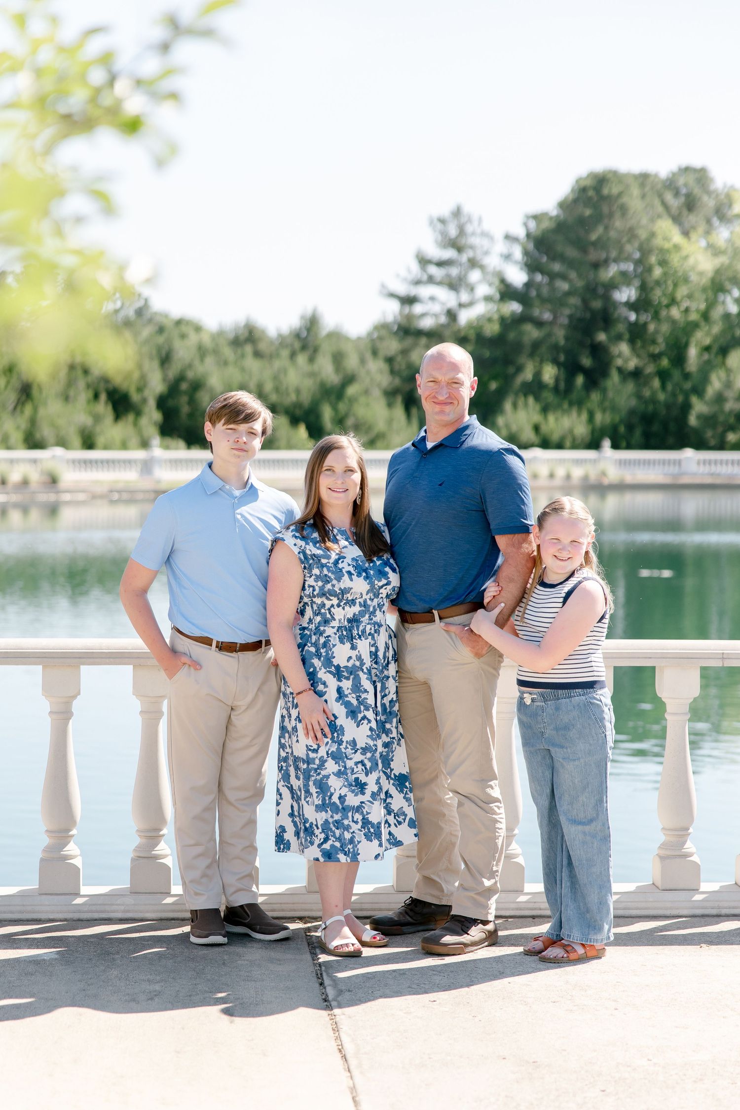 Family of four poses together on stone balcony overlooking peaceful lake on sunny summer day.