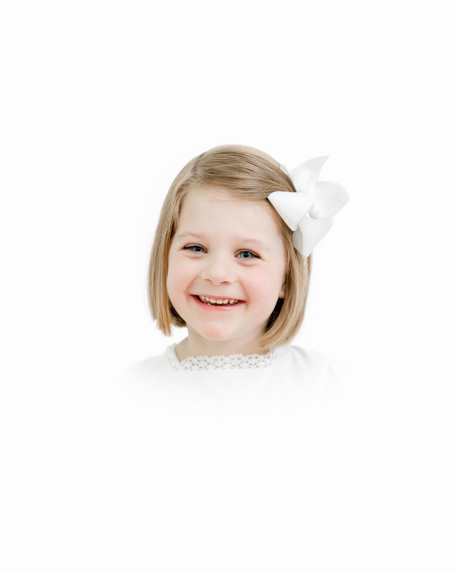 A young child with short blonde hair wearing a white lace dress and bow smiles brightly.