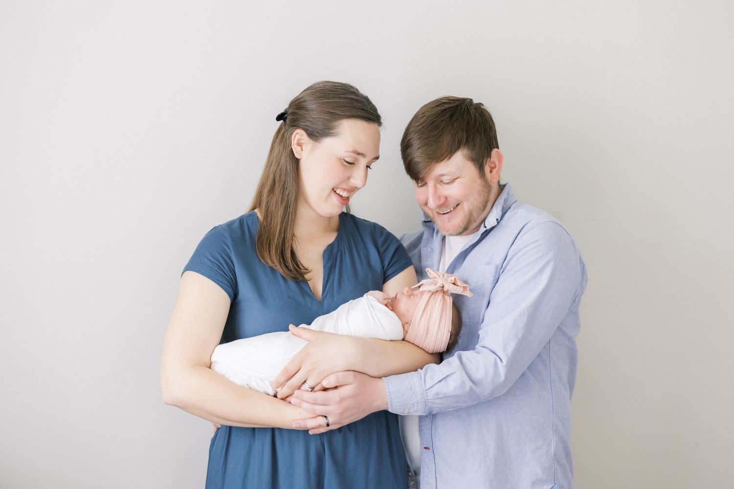 A couple stands together against a gray wall sharing a loving moment with their newborn baby wrapped in white.