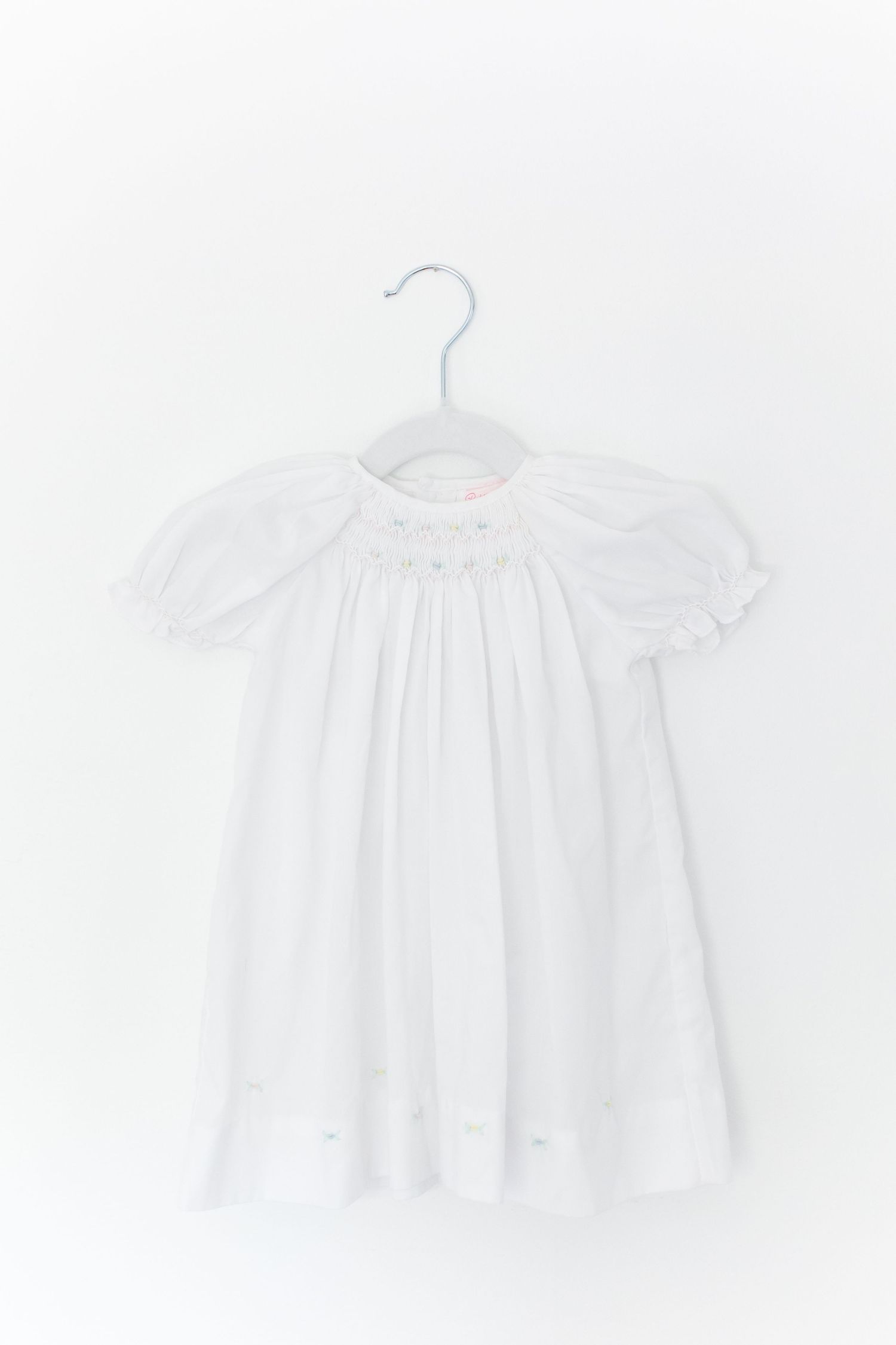 Delicate white smocked baby dress with puff sleeves and pastel floral embroidery hanging on grey hanger.