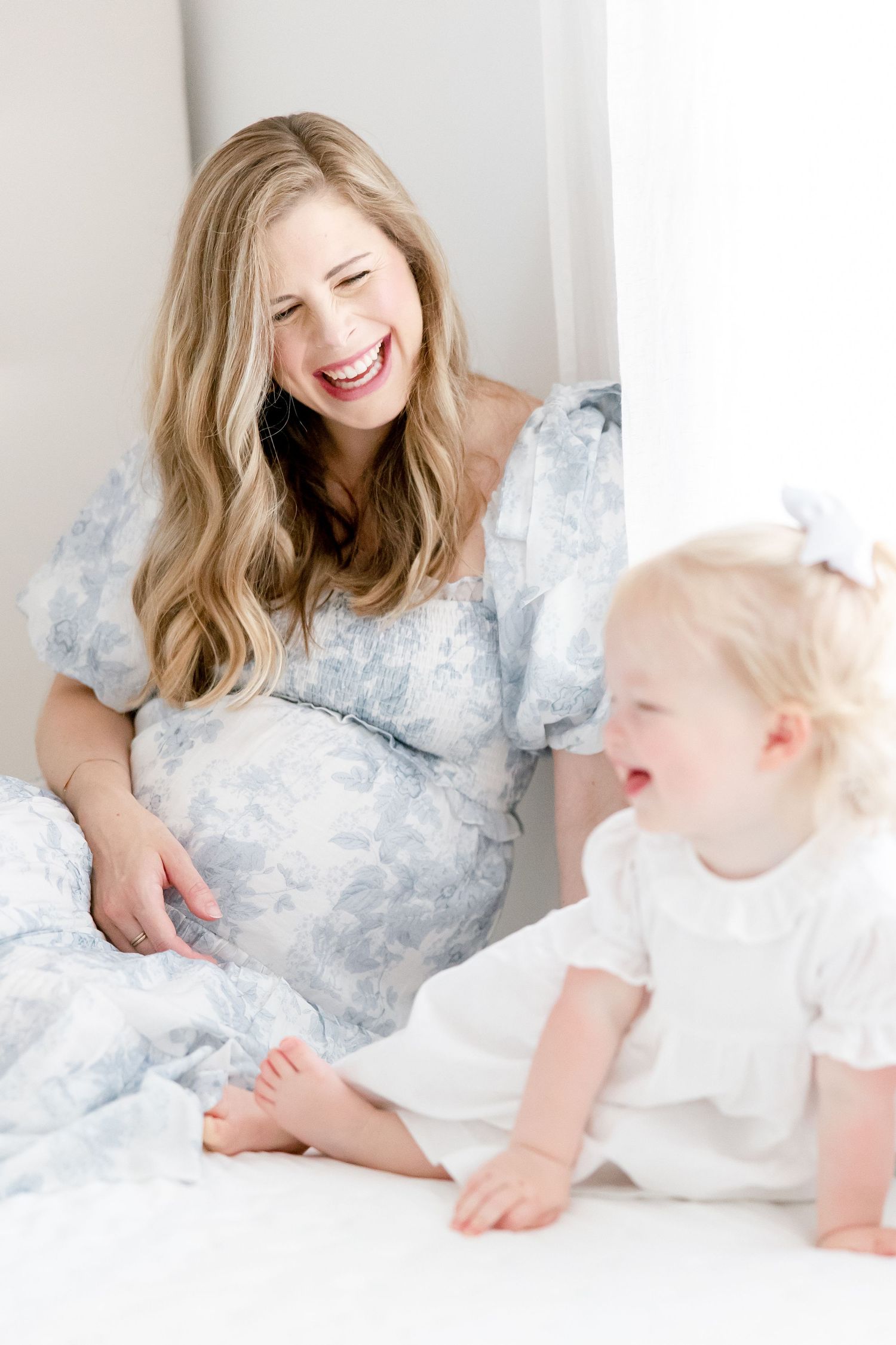 Joyful moment shared between mother and child on white bedding in bright natural light.