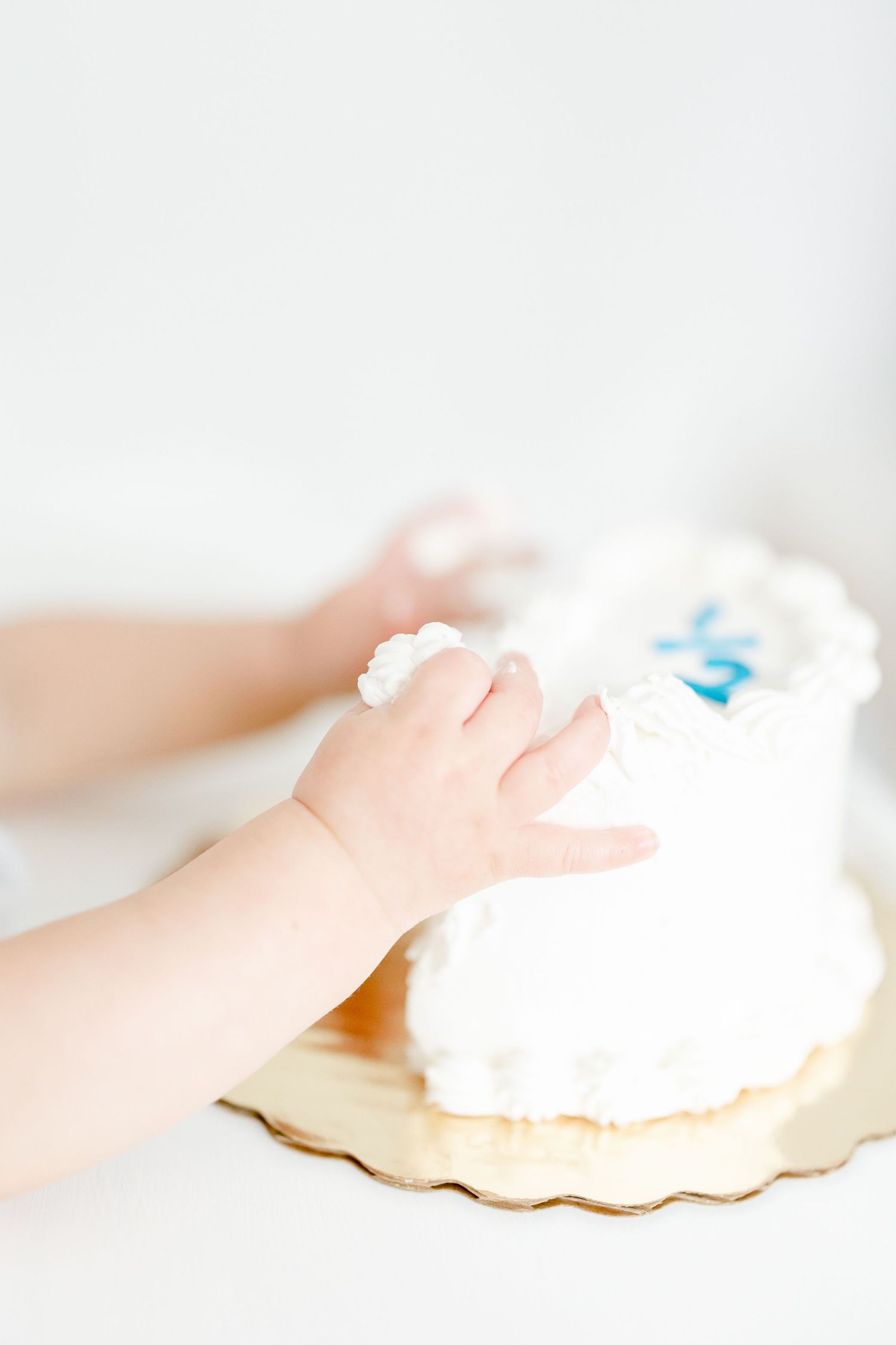 Small baby hand reaches for white frosted cake in soft, bright lighting.