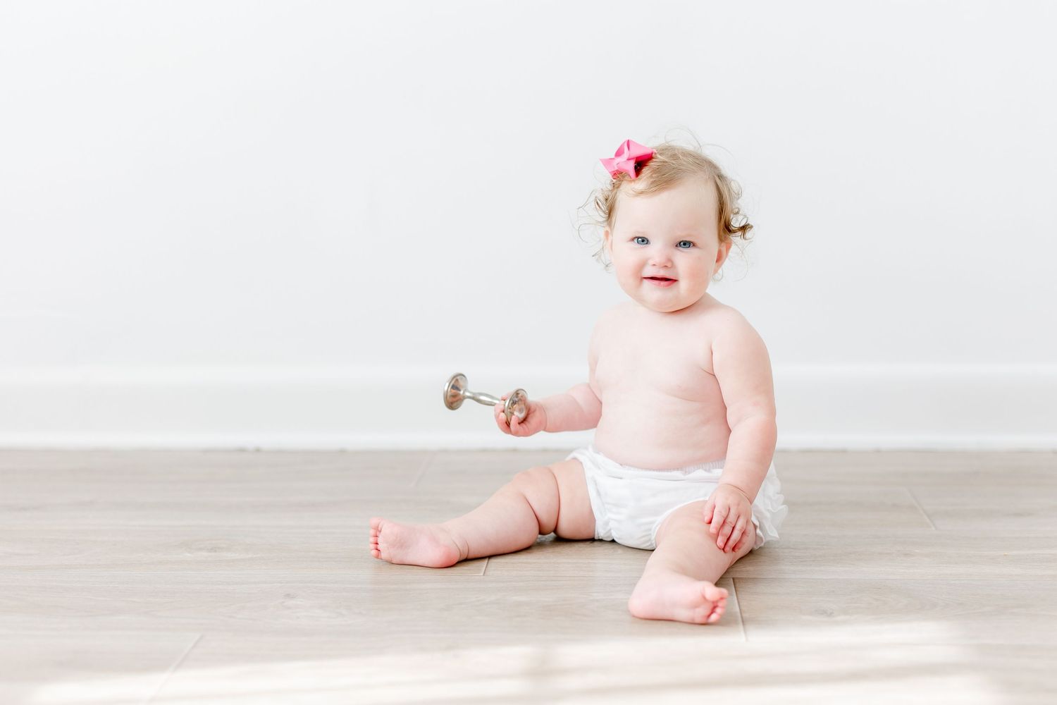 Baby in pink dress sitting on light wooden floor with small pink bow hair accessory.