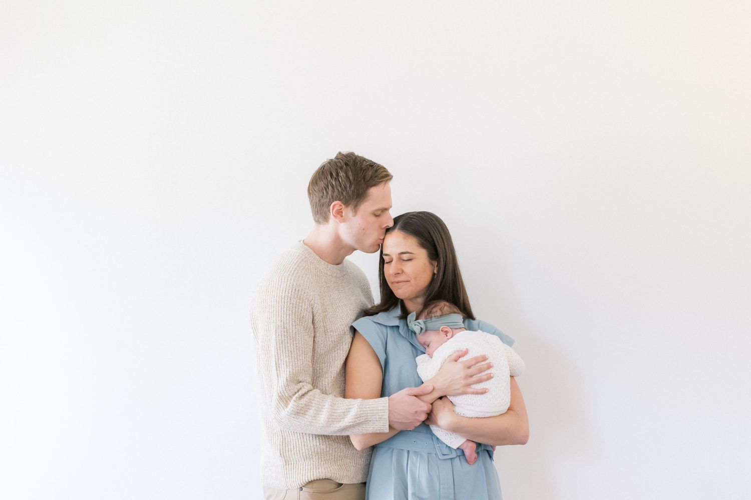 A tender couple embraces against a white wall while sharing an intimate moment together.
