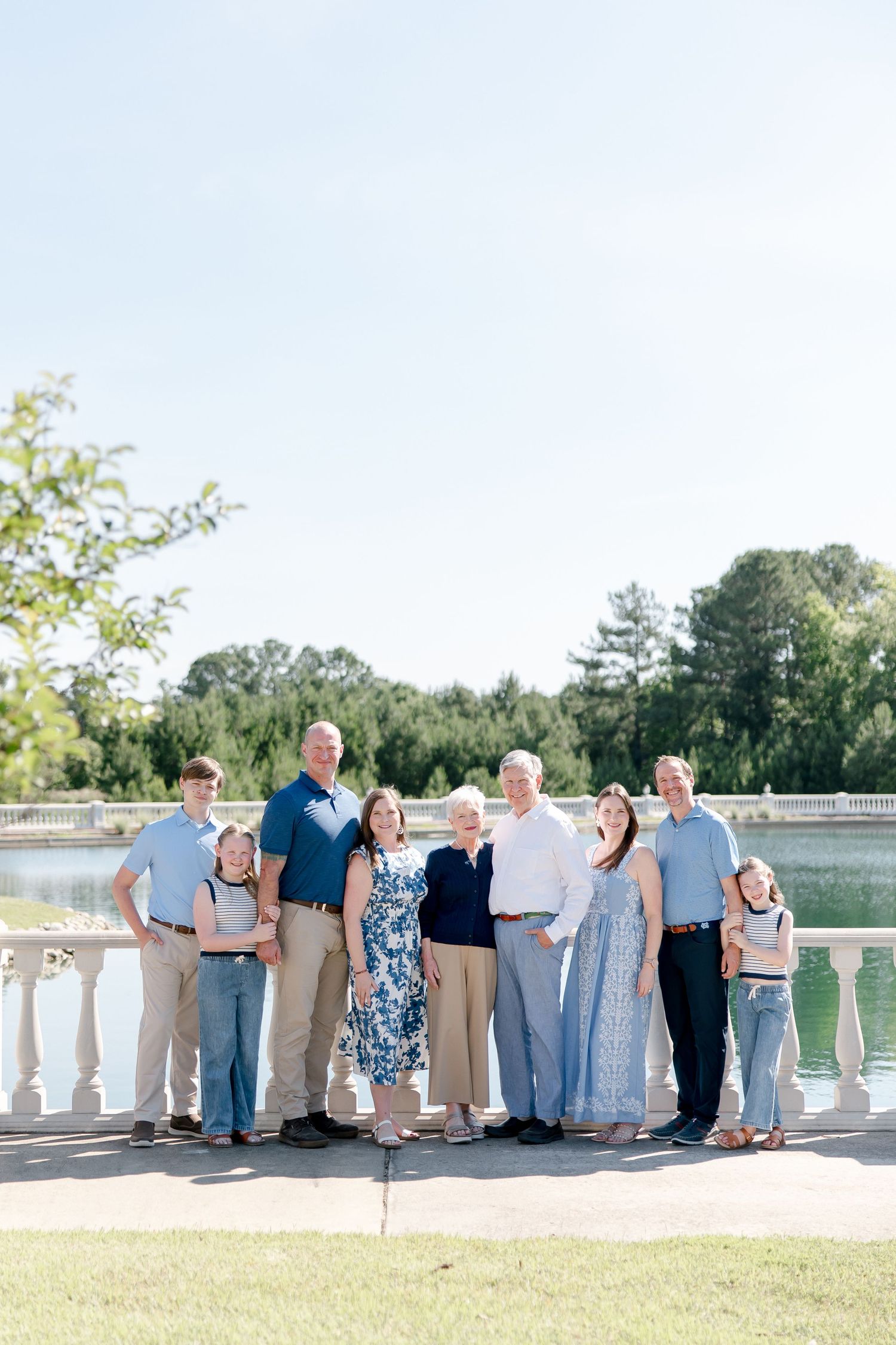 A multi-generational family stands together on a stone balcony overlooking a serene lake on a sunny summer day.
