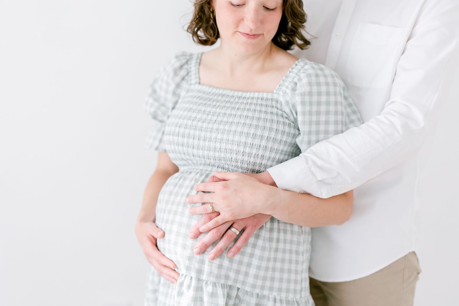 A person wearing a gray and white gingham dress holds their pregnant belly while standing against a white wall.