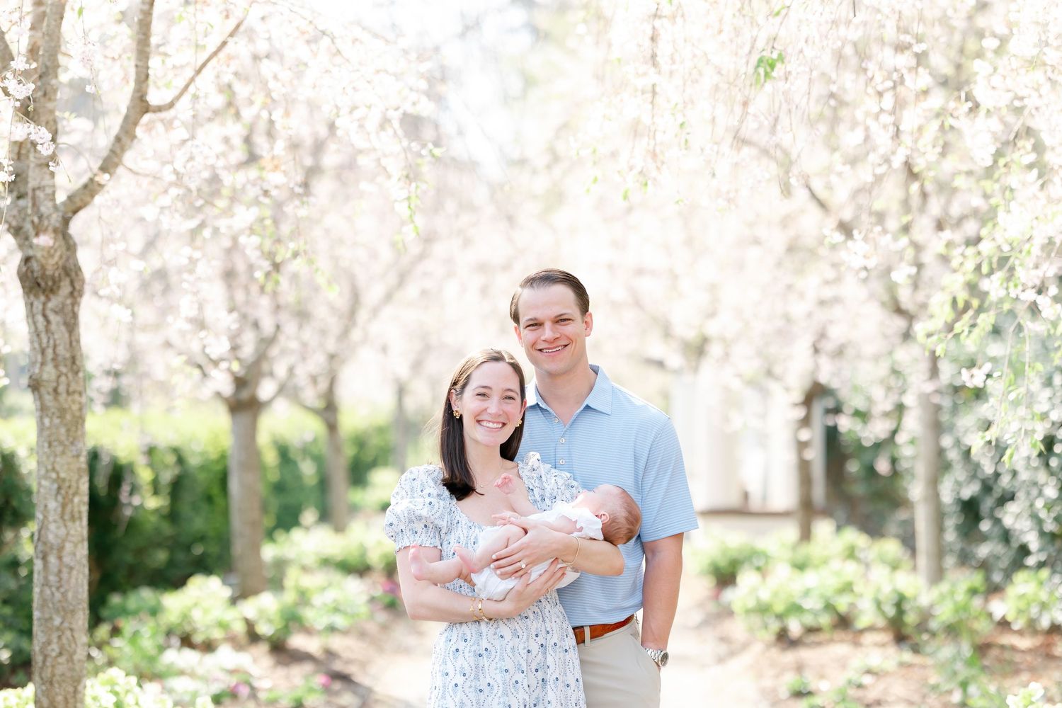 A smiling couple holds their newborn baby among blooming cherry blossom trees on a sunlit garden path.