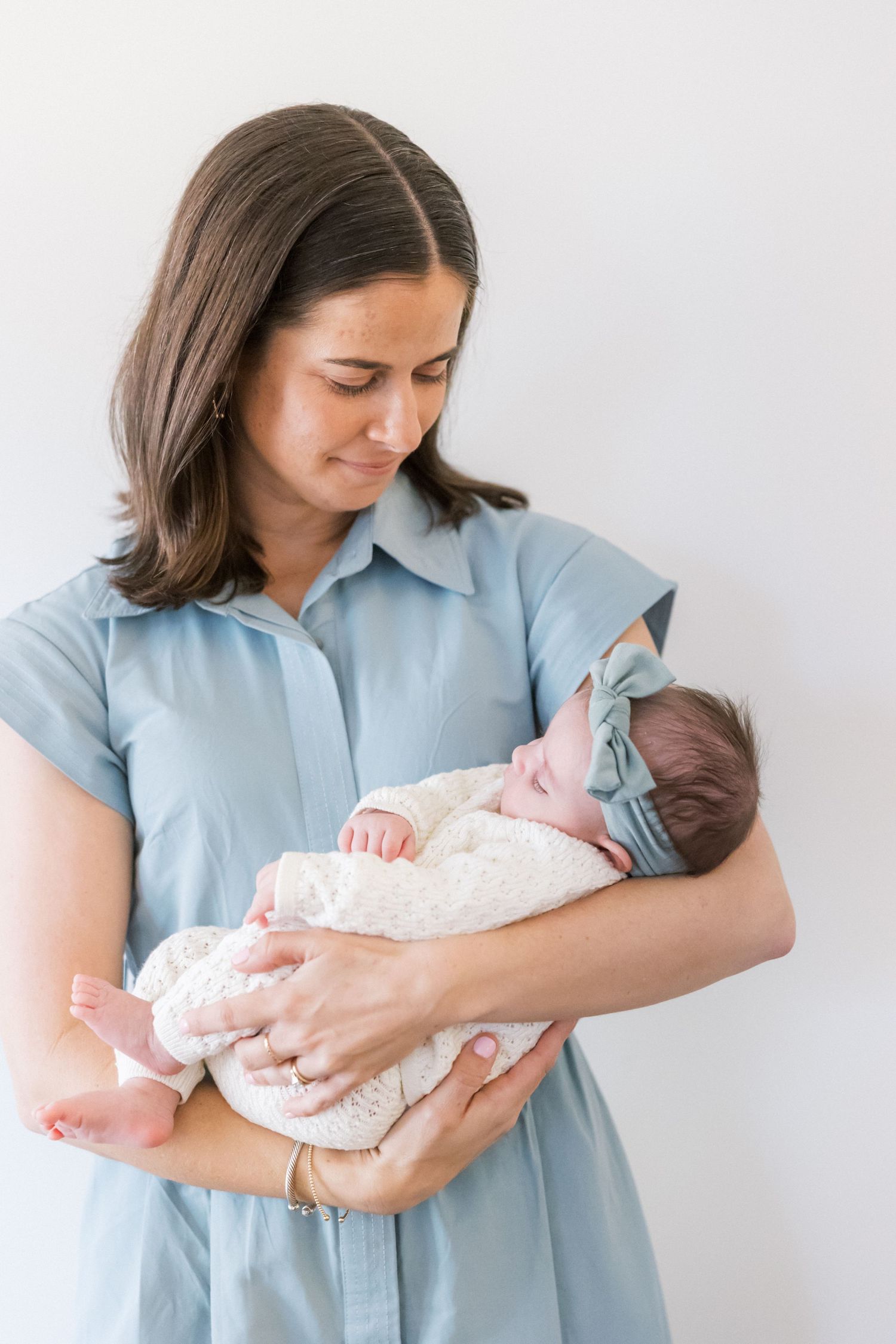 Person in light blue dress holds infant wearing cream knit outfit and mint headband against white background.