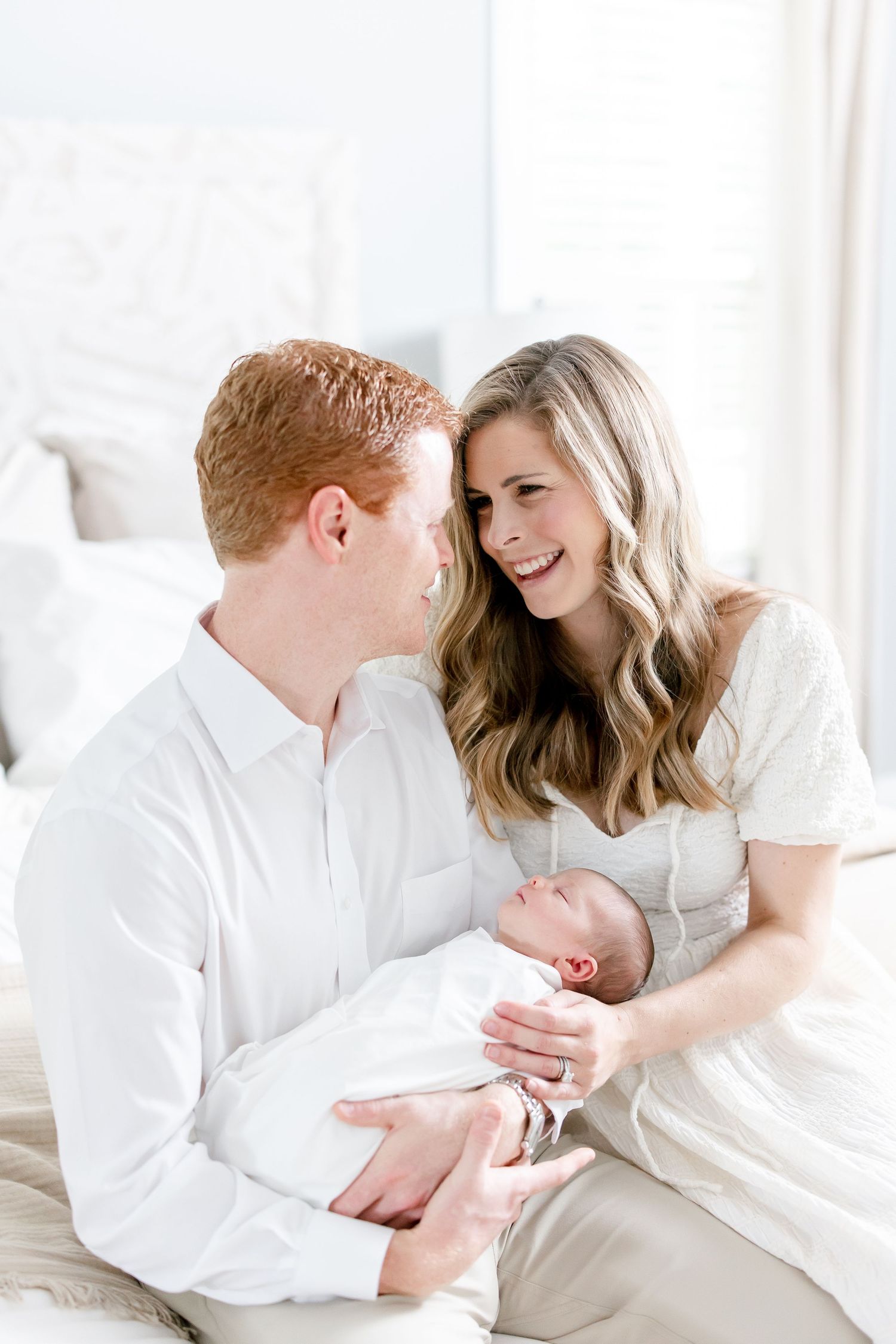 A couple dressed in white sits on a bed sharing an intimate moment while holding their newborn baby.