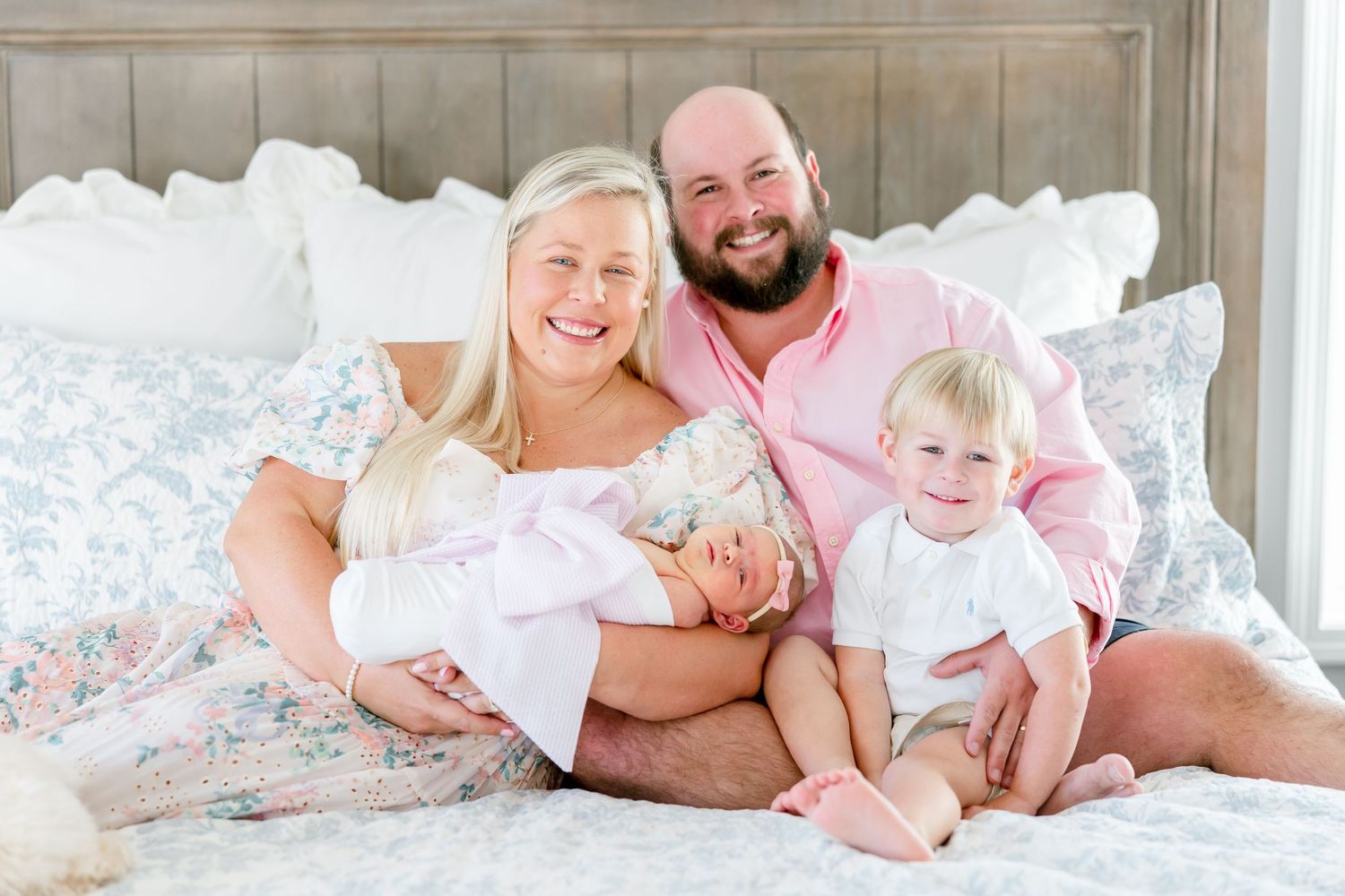 A group relaxes together on a white bed with soft pillows and floral bedding.
