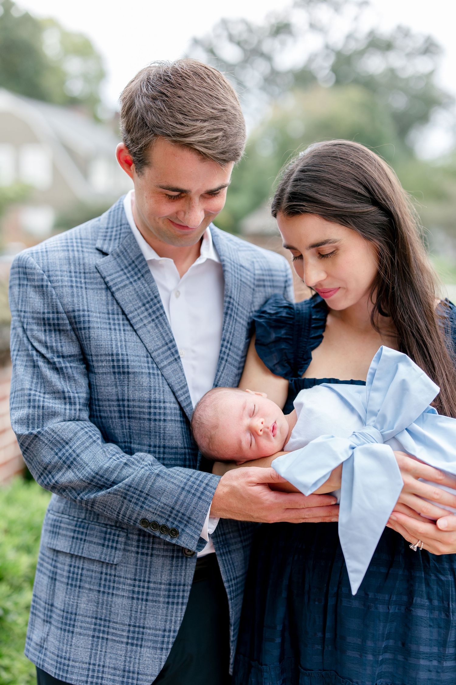A couple in formal attire holds and gazes at their newborn baby while standing outdoors.
