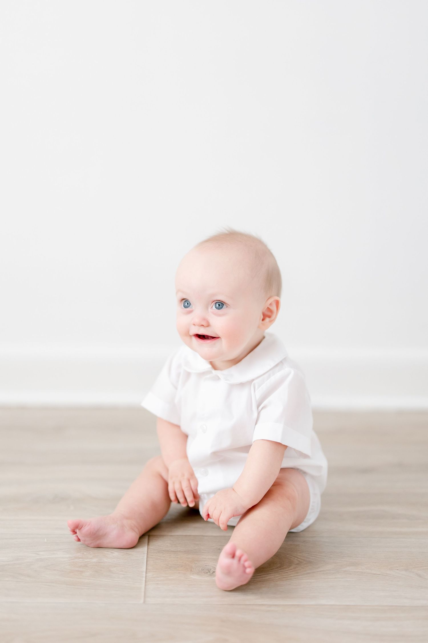 Baby in white outfit sits on wooden floor against white wall, showing a happy expression.