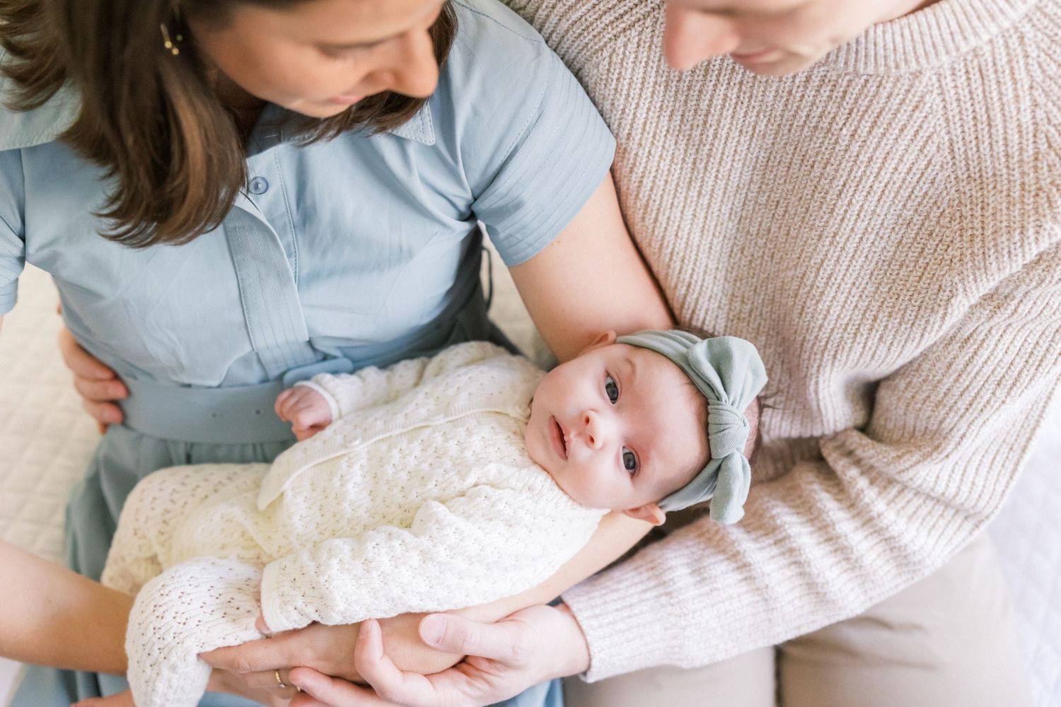 Baby wearing cream knit outfit and blue bow headband lies in arms with cream sweater and blue shirt visible.