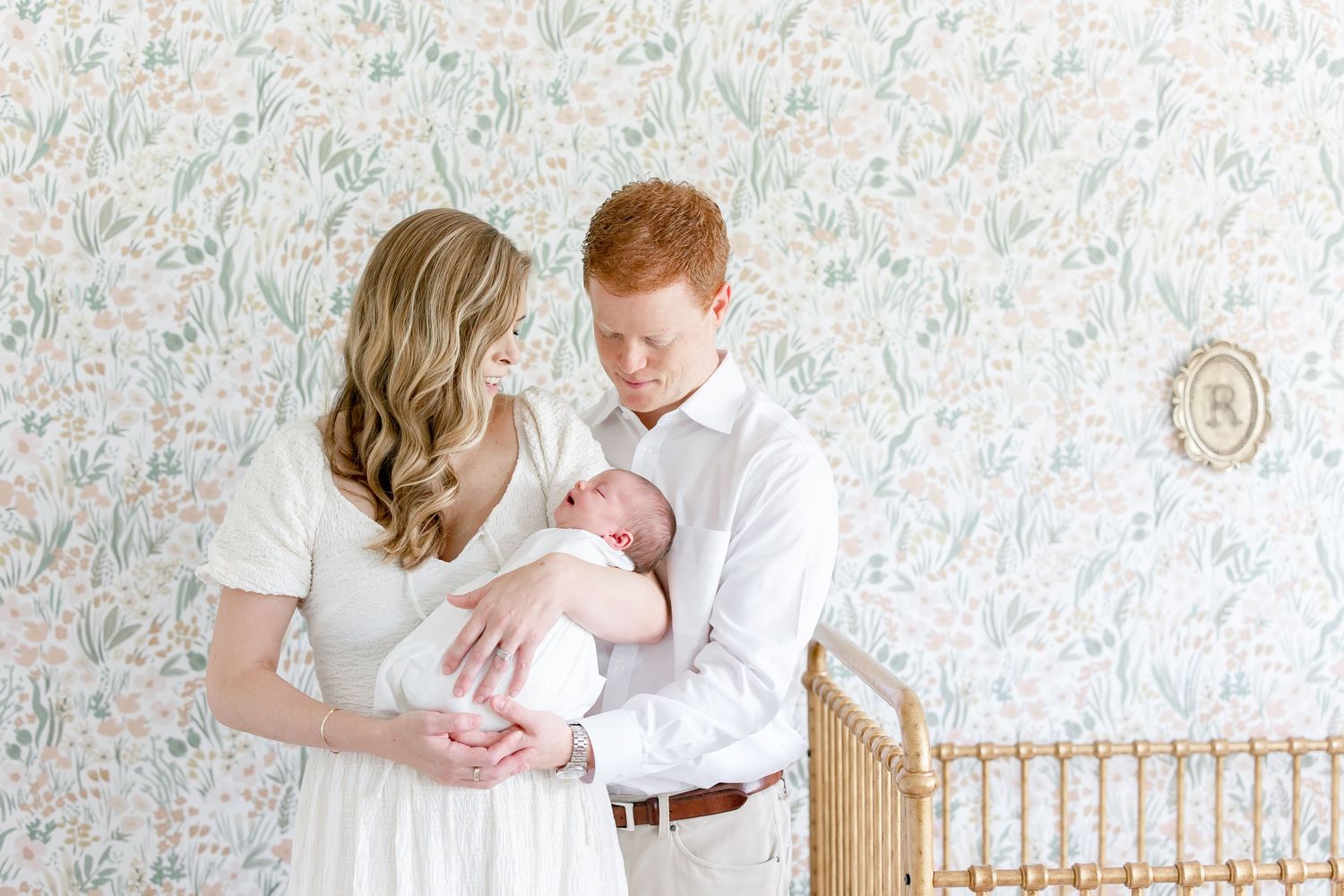 A nursery with parents and baby standing by a white crib near a floral wallpapered wall.