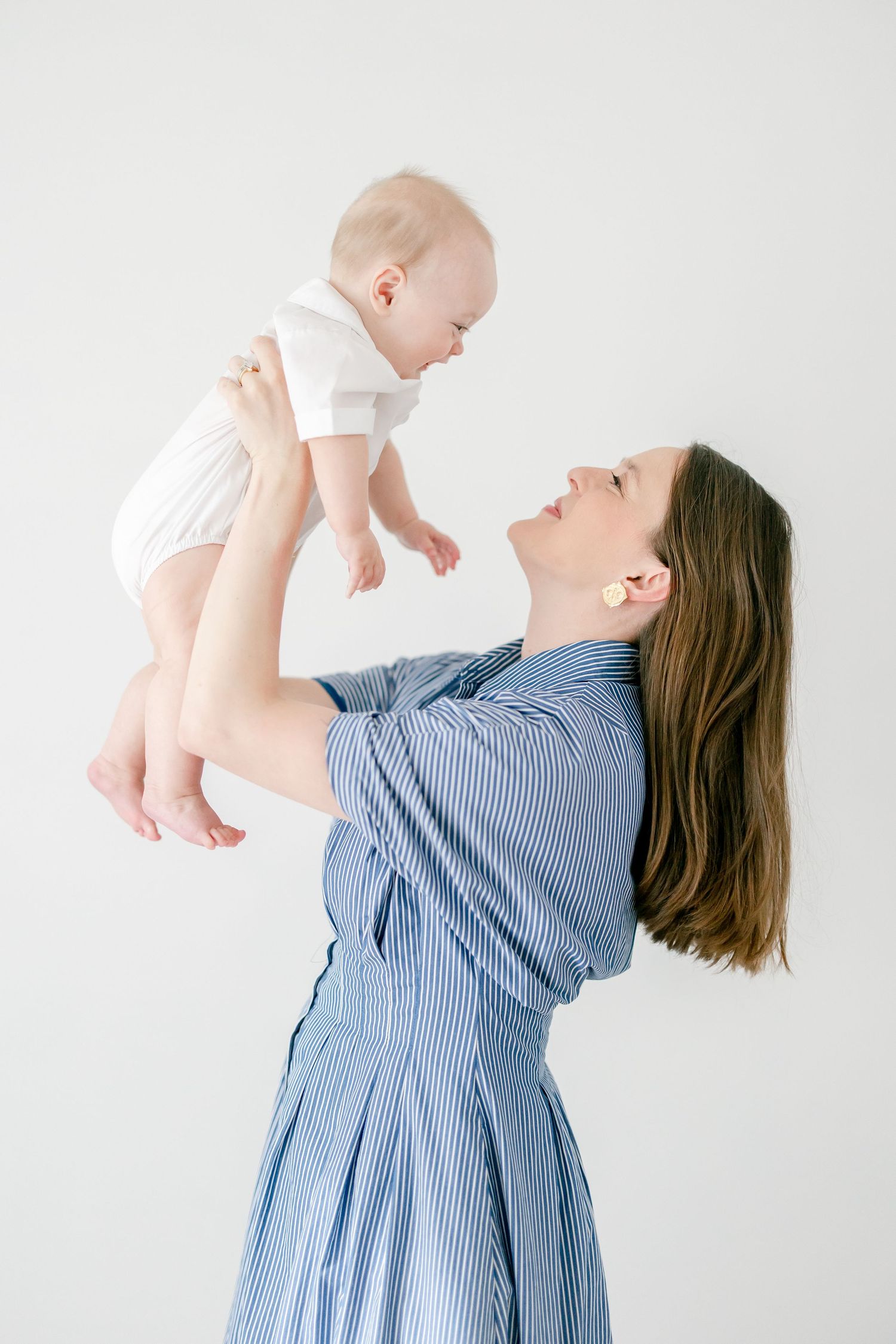 A woman in a blue striped dress lifts and plays with a baby wearing white against a light background.