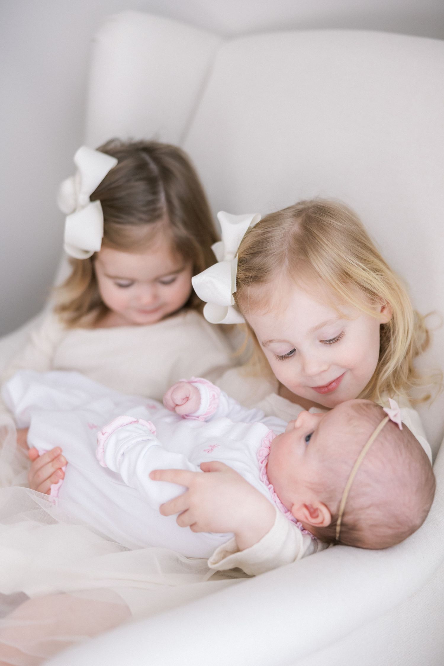 Two young children in white bows sit together on a light-colored couch while holding and gazing at a small baby.
