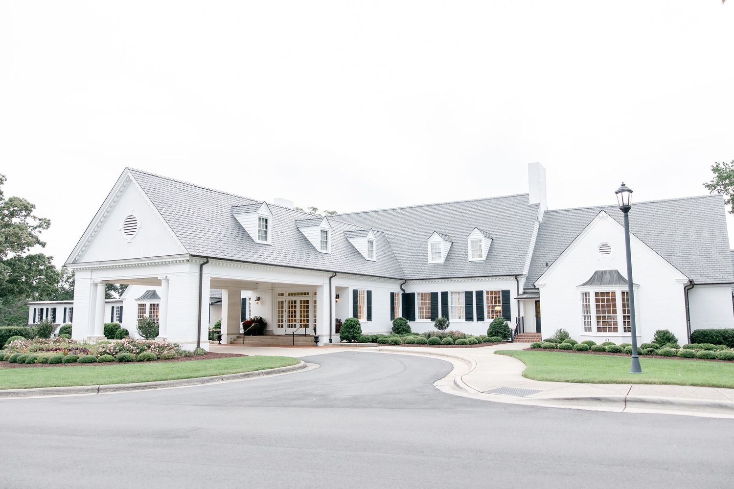 White luxury farmhouse exterior with dormer windows, covered porch and wrap-around walkway creates elegant curb appeal.