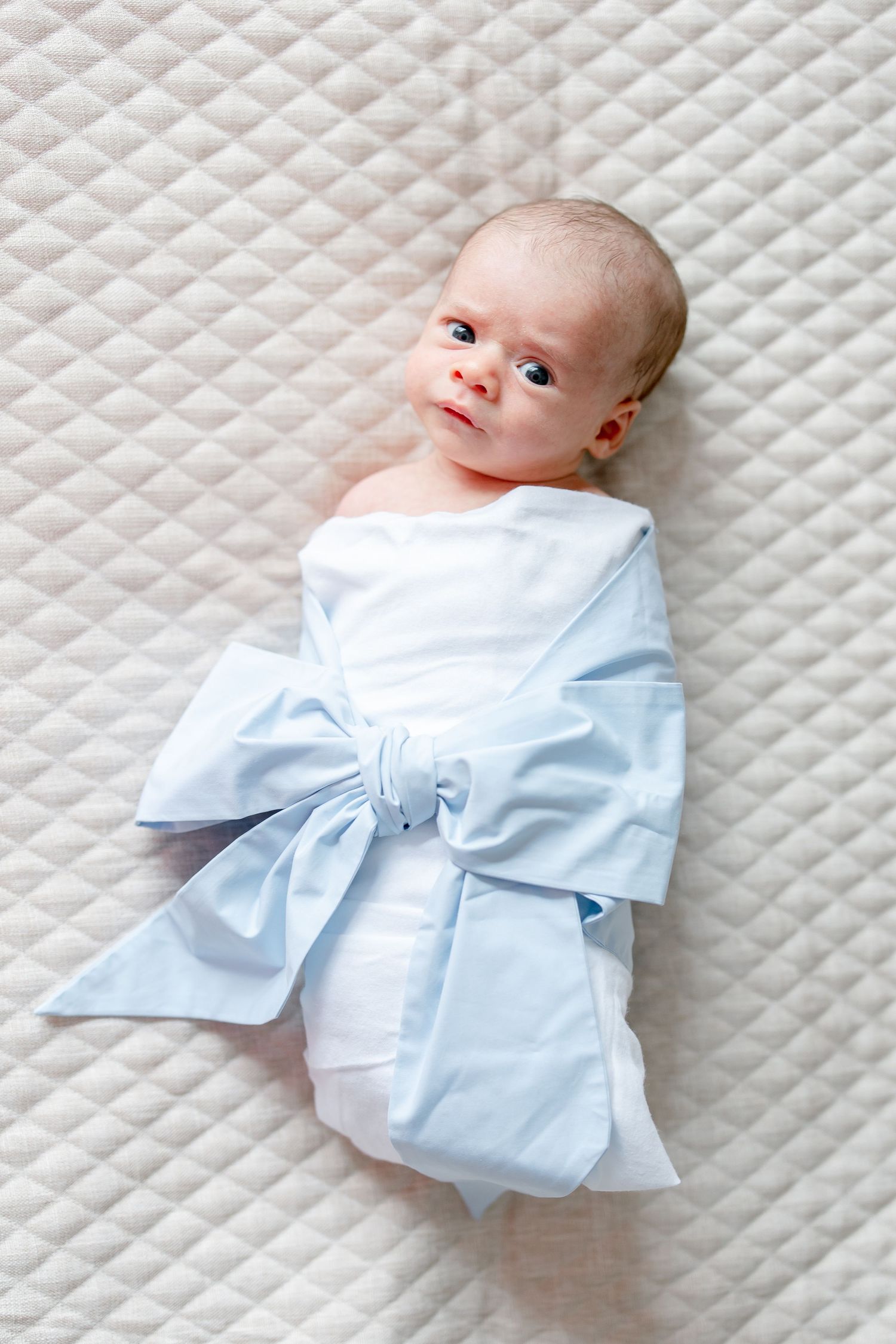 Baby in light blue swaddle wrap lying on quilted white surface looking sideways.