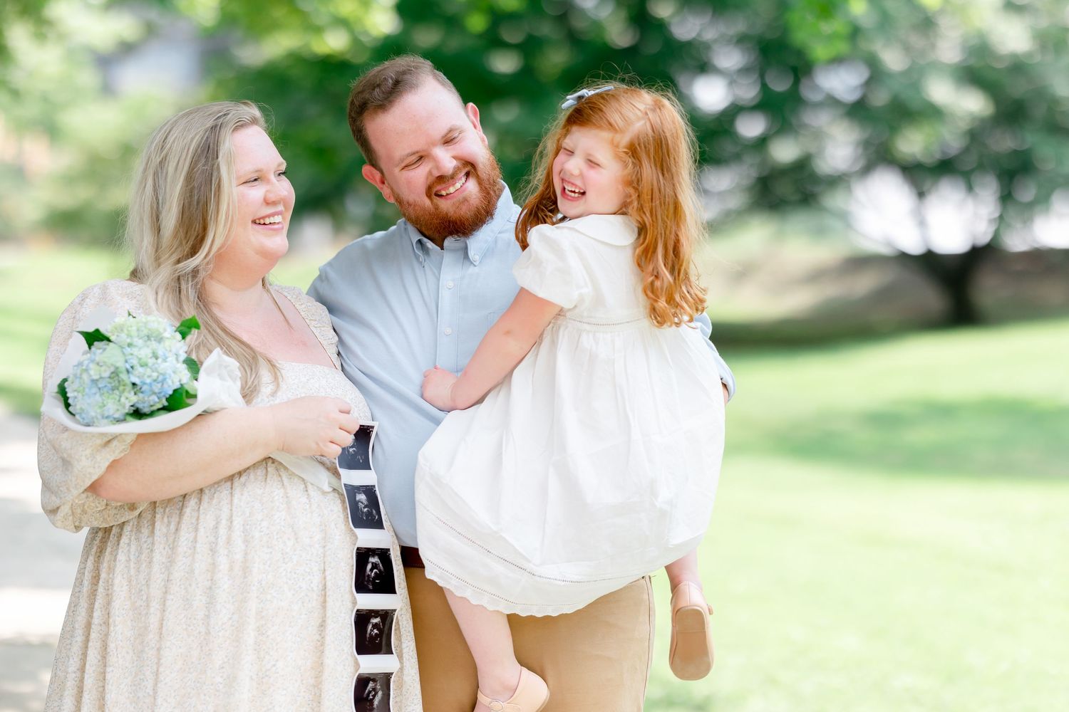 Smiling pregnant woman holding flowers stands with bearded man carrying laughing toddler girl outdoors.