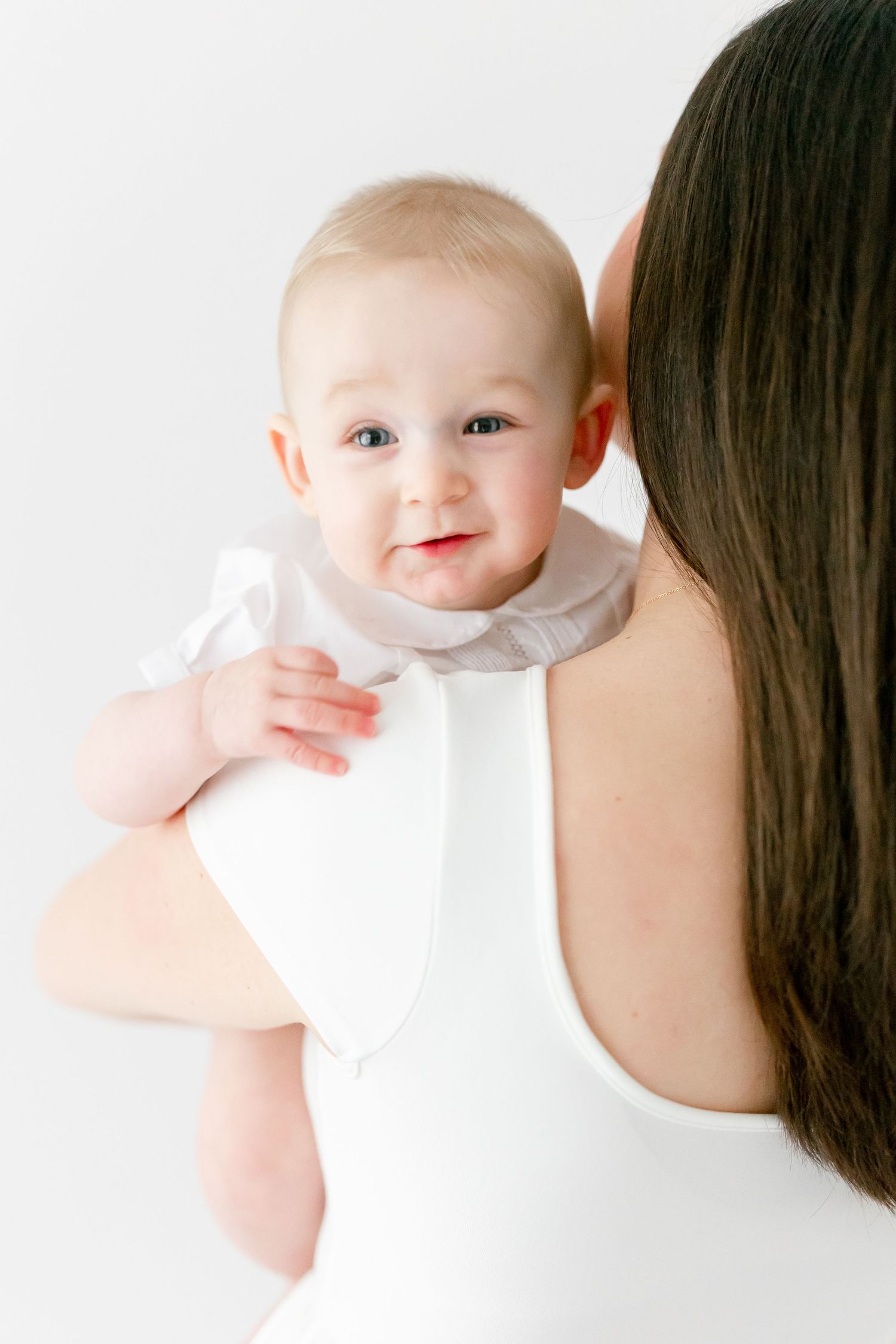 Smiling baby with light hair in a white outfit looks at the camera while resting on the shoulder of a woman with long dark hair.