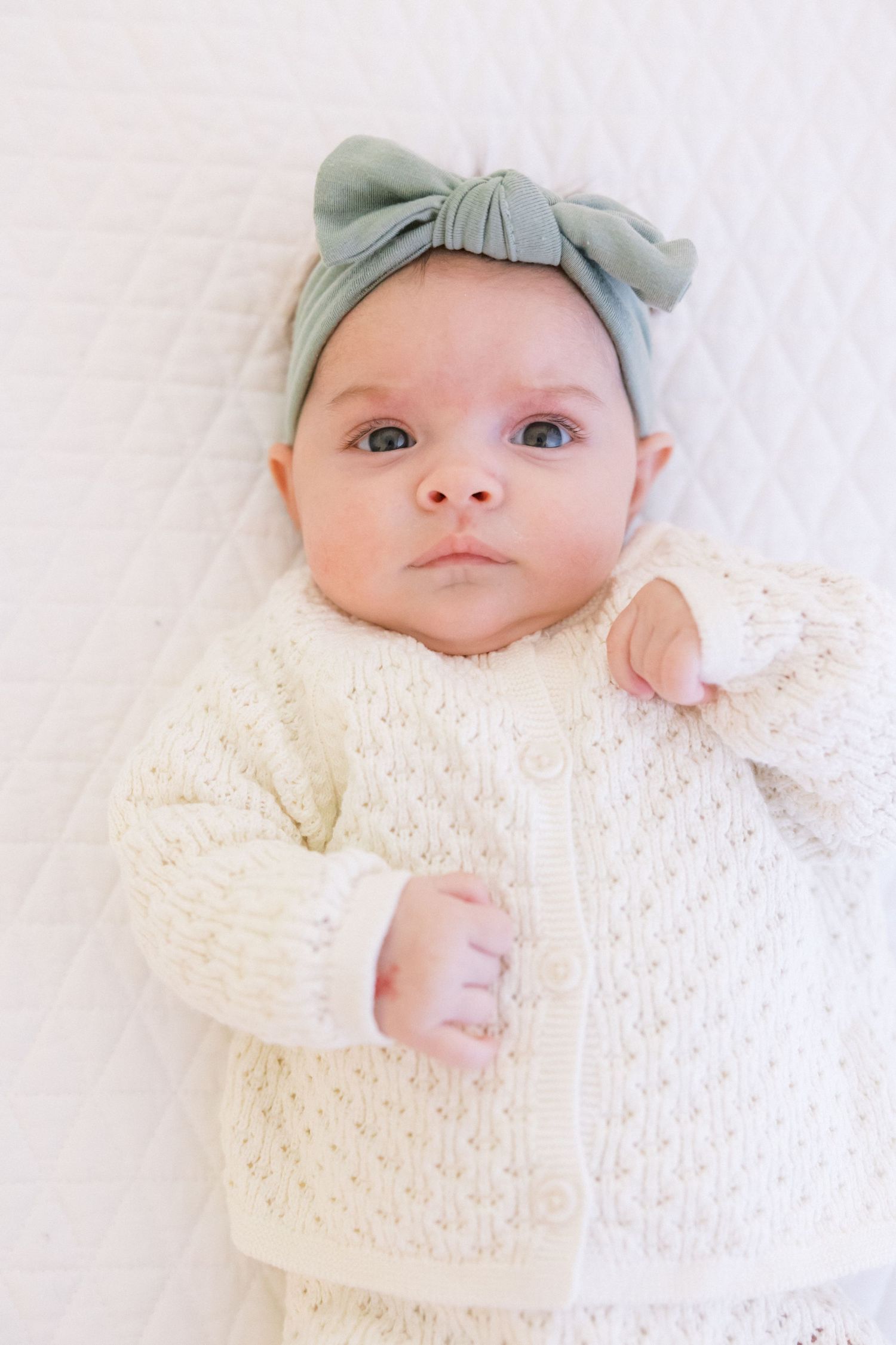 Sweet newborn baby wearing a mint green cap lies swaddled in soft cream knit blanket.