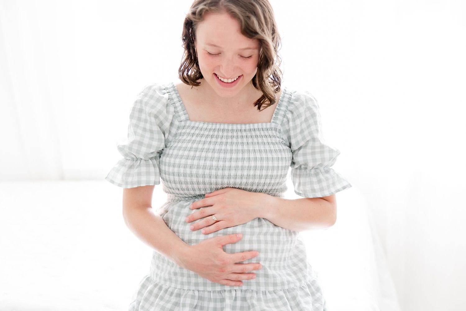 Pregnant woman in a light gingham dress holding her baby bump while standing against a white background.