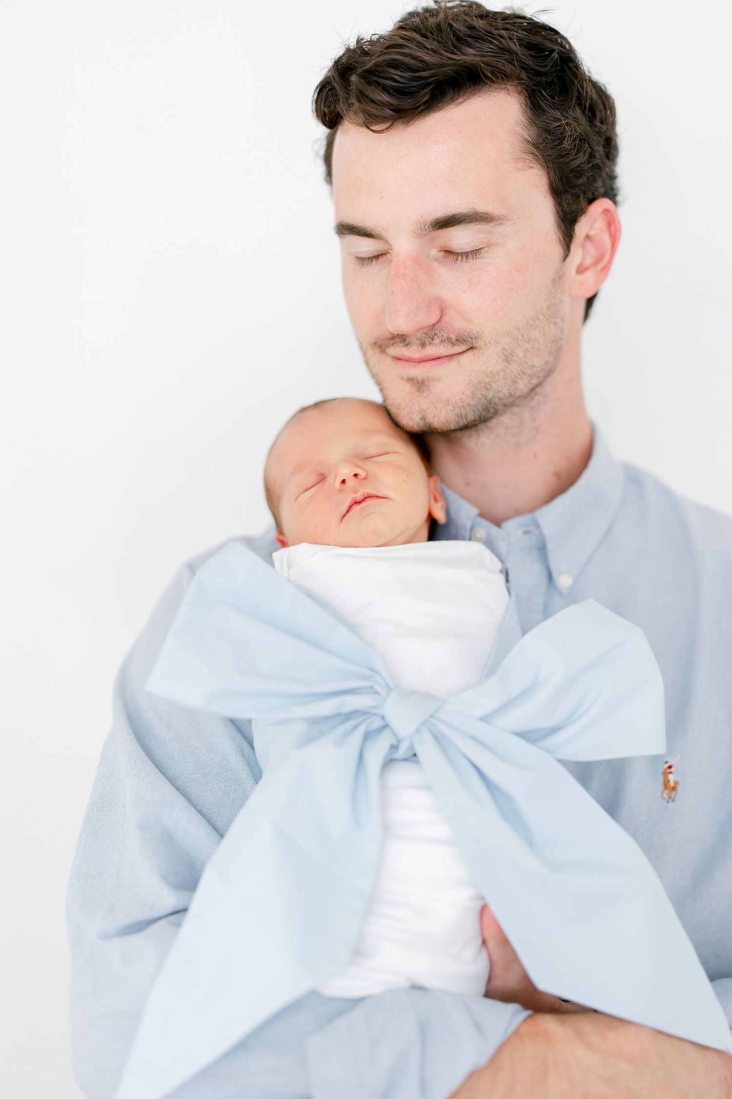 A tender moment shows a parent in a light blue shirt holding their sleeping newborn baby wrapped in a blue bow swaddle.