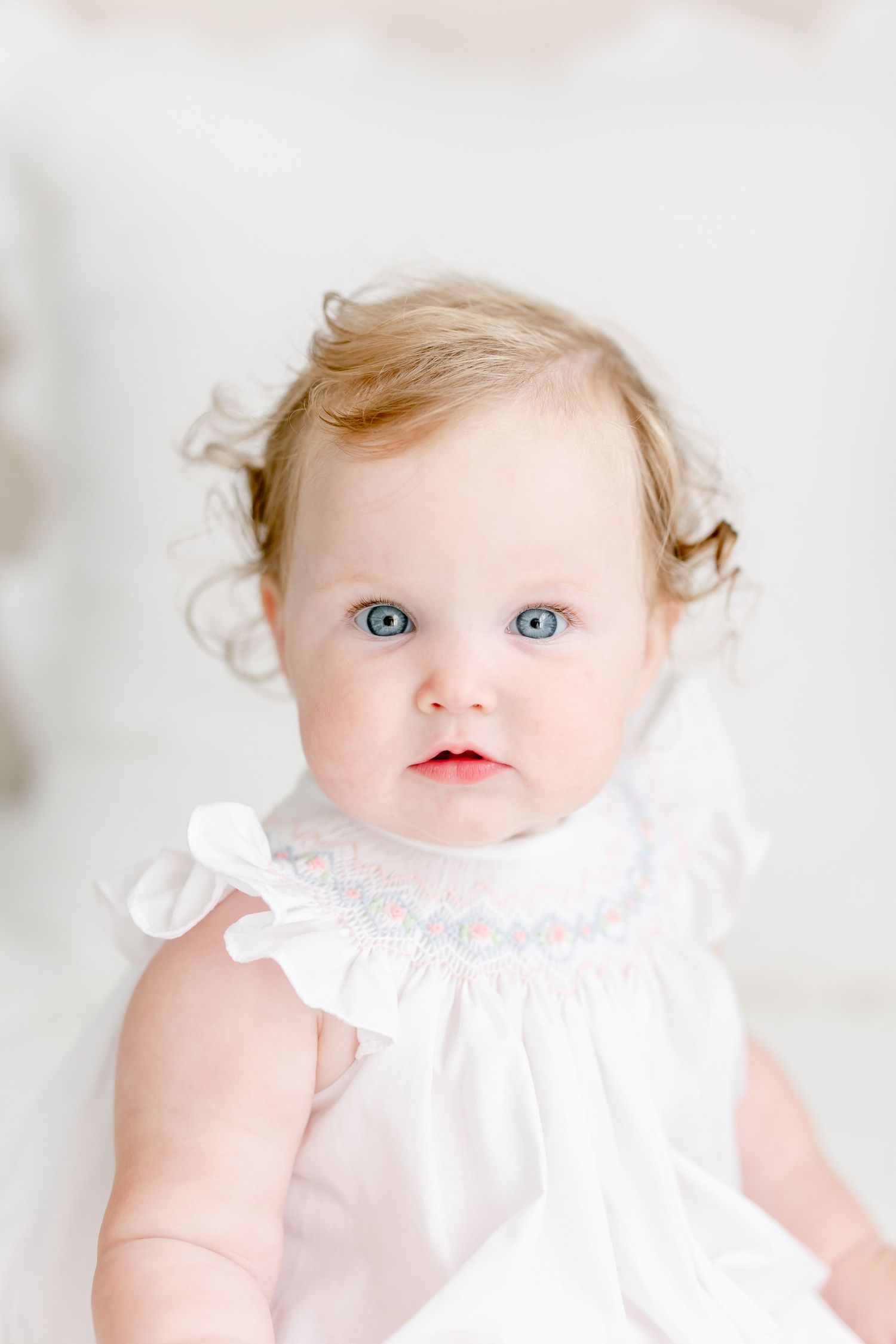 Baby wearing a white smocked dress with ruffles and blonde curls gazes at camera with bright blue eyes.
