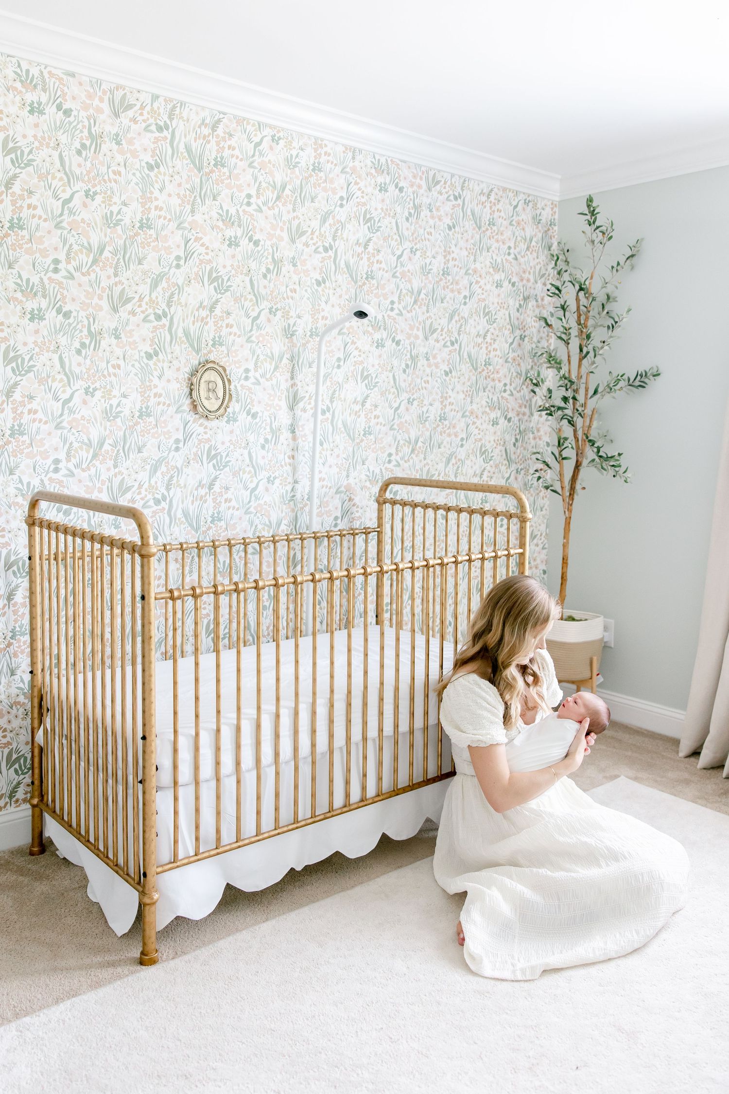 A nursery with floral wallpaper showcases a gold vintage-style crib and decorative tree beside it.