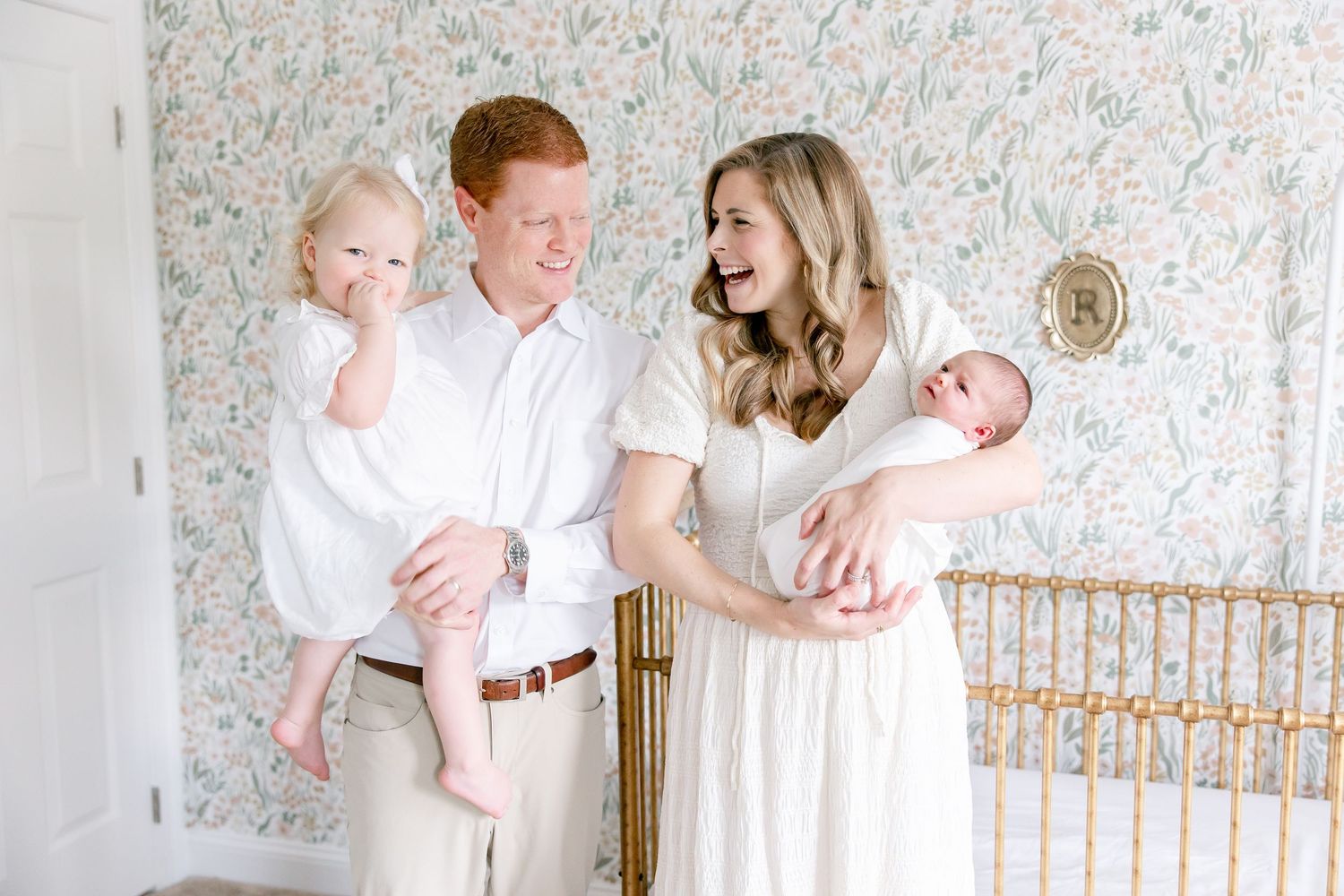 Parents in white outfits pose with their newborn baby in a bright nursery setting with floral wallpaper.