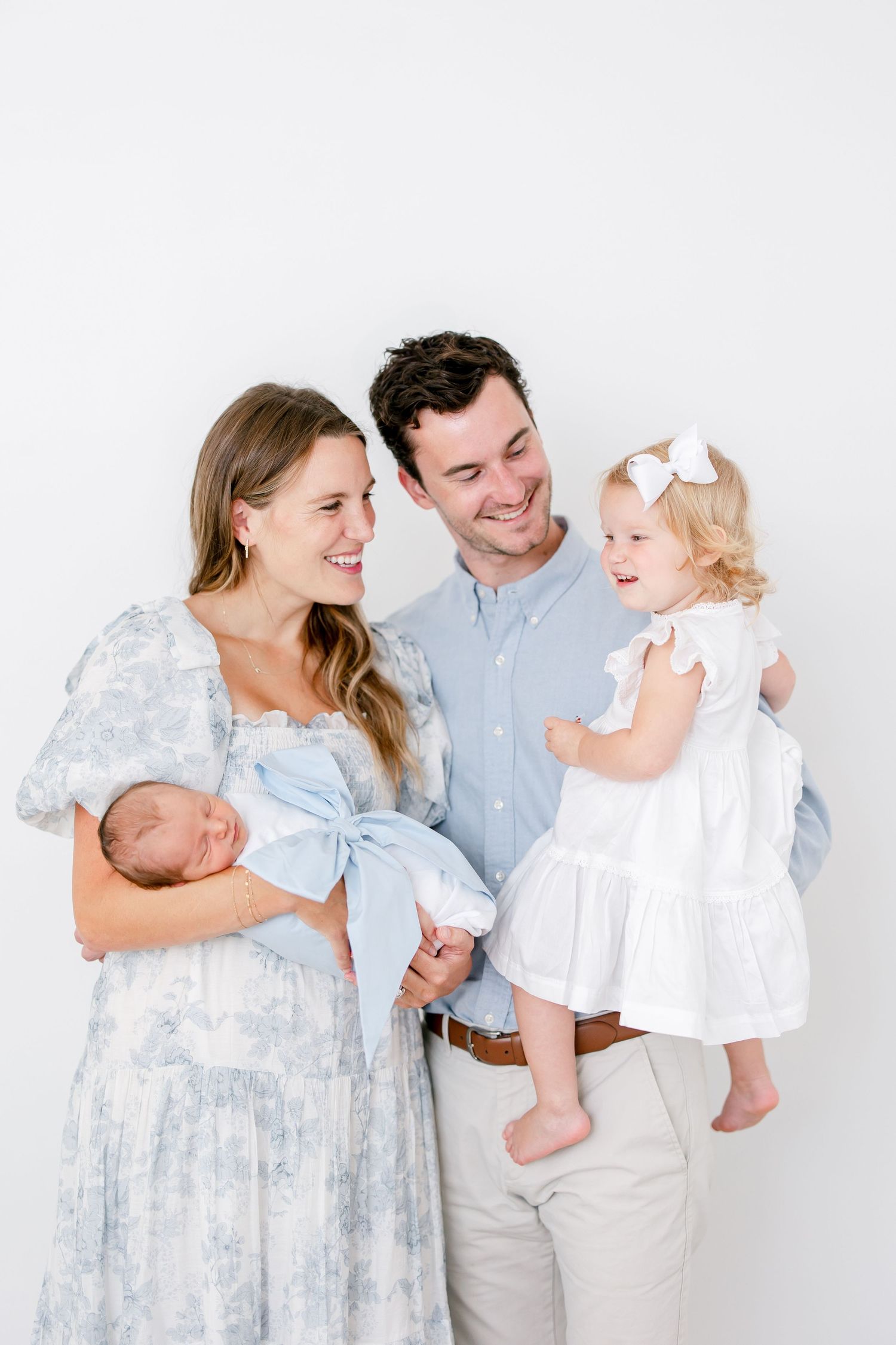 Family portrait with parents and two children against white background wearing light blue and white clothing.
