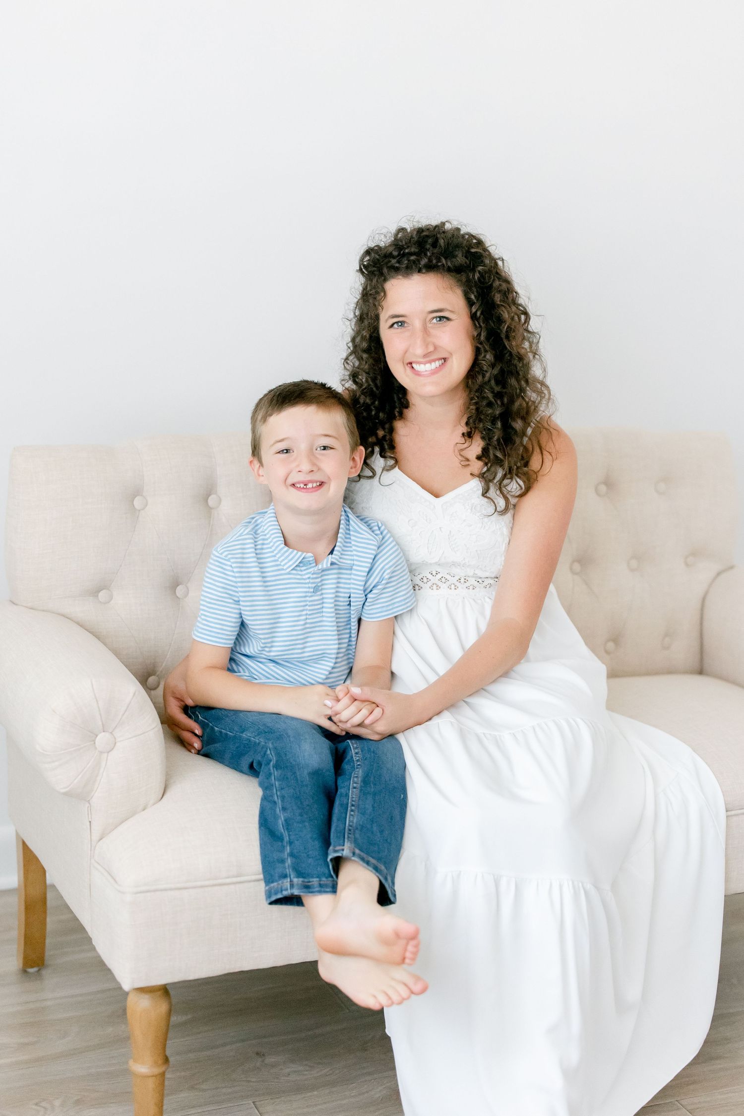 Mother and child wearing white and blue clothes share playful moments on a light-colored couch.