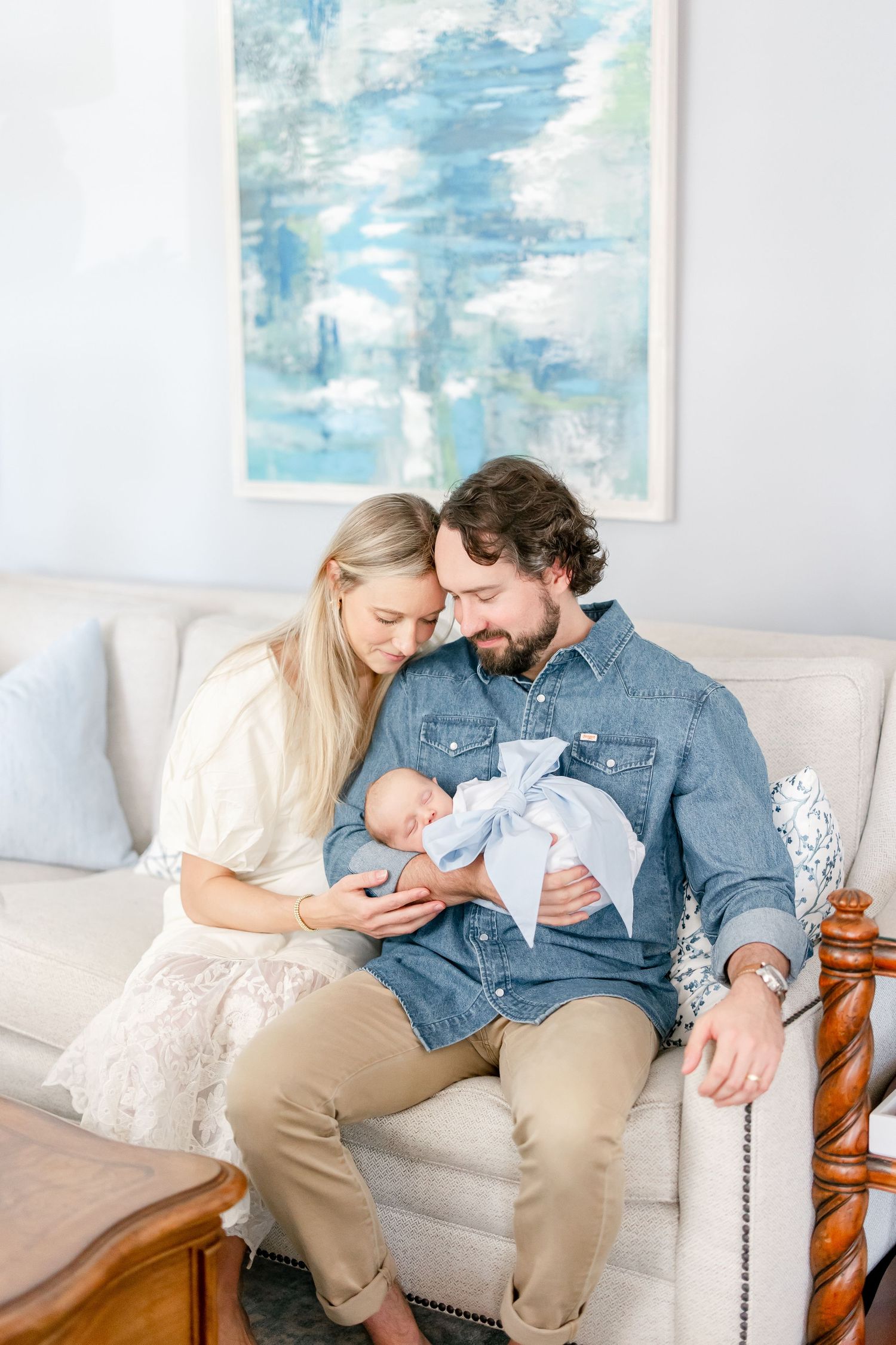 Parents cuddling with newborn baby on white couch with blue abstract art on wall.