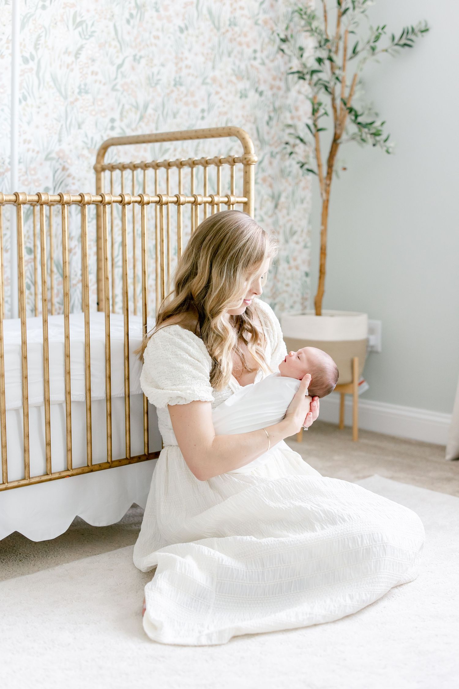 Nursery scene with mother in white dress holding baby near gold crib with floral wallpaper.