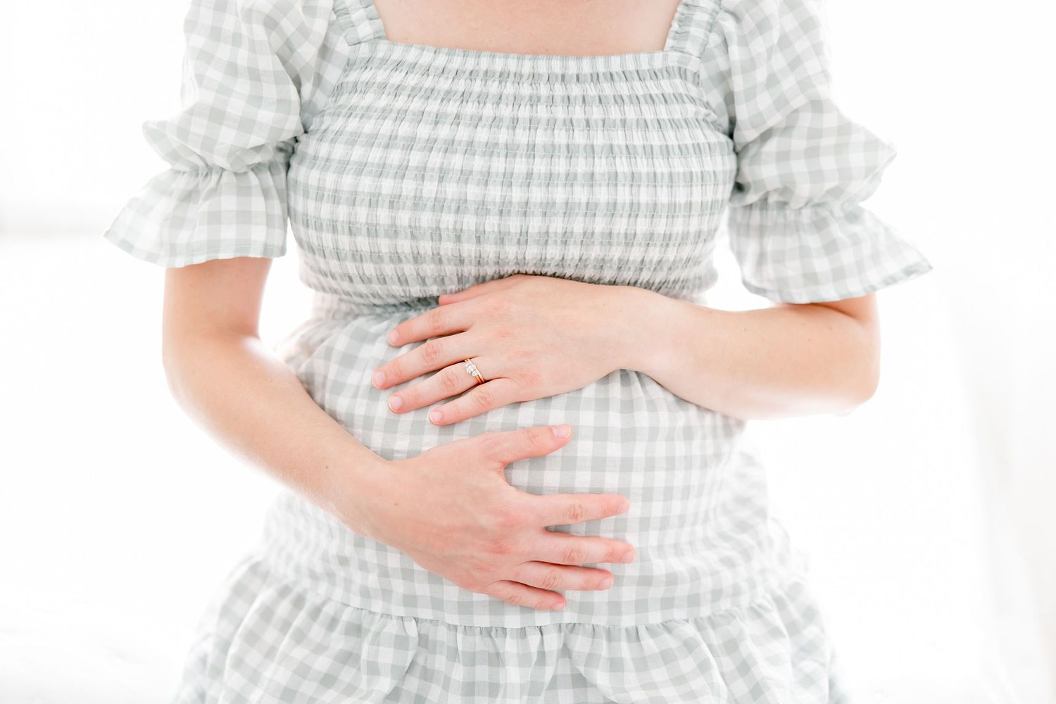 A person wearing a light gingham checkered dress holds their stomach with both hands against a white background.