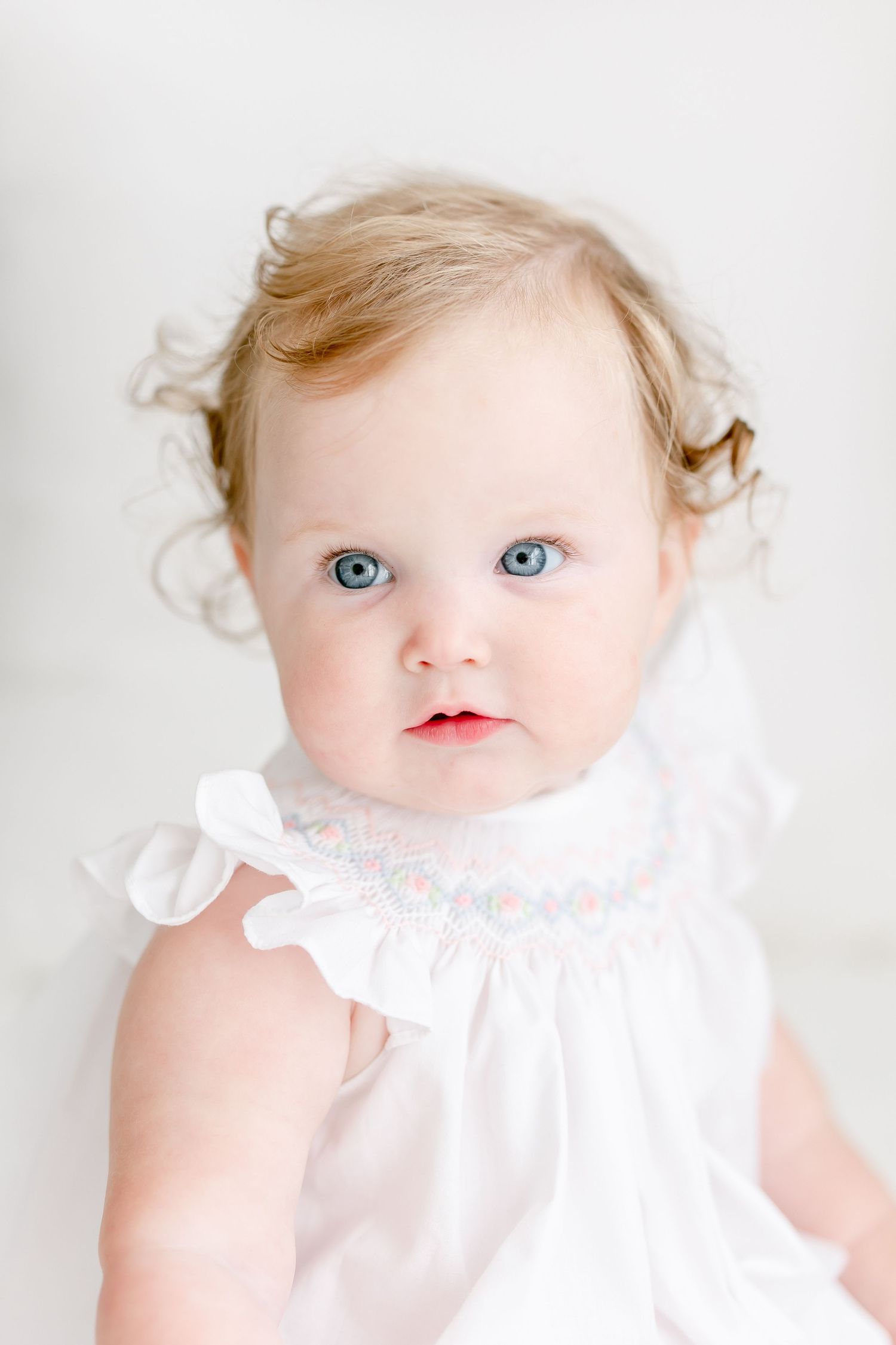 Baby wearing white dress against light background with bright blue eyes and blonde curly hair looking up at camera.