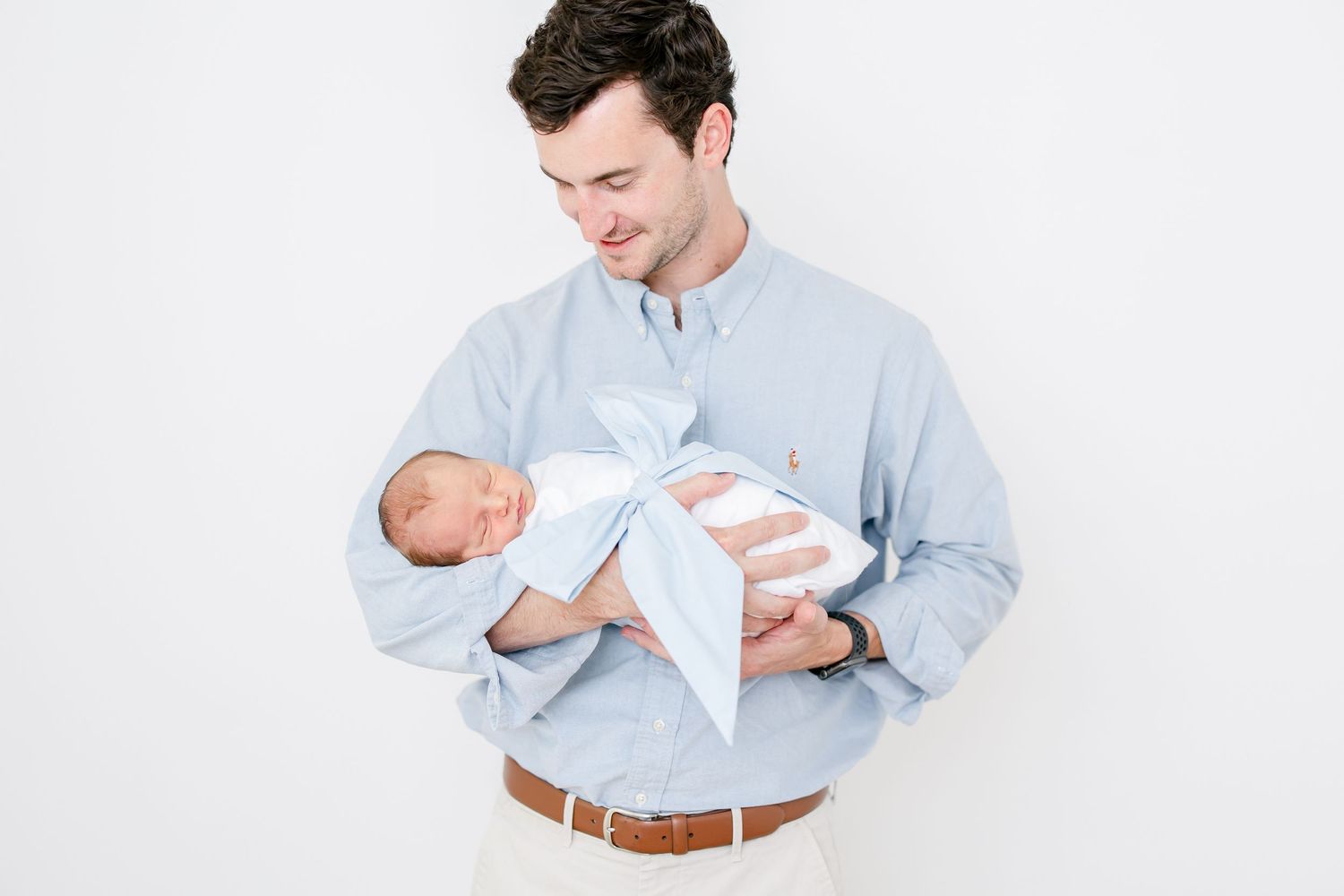 A person in a light blue button-down shirt tenderly holds a sleeping newborn baby against a white background.